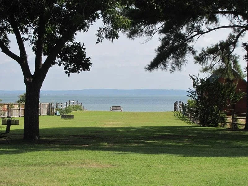 View of a grassy lawn leading to a wooden fence and a large body of water, with trees providing shade on the lawn. A bench is visible near the water's edge and there is a house on the right side. Hill Country Texas lake cabin rentals.