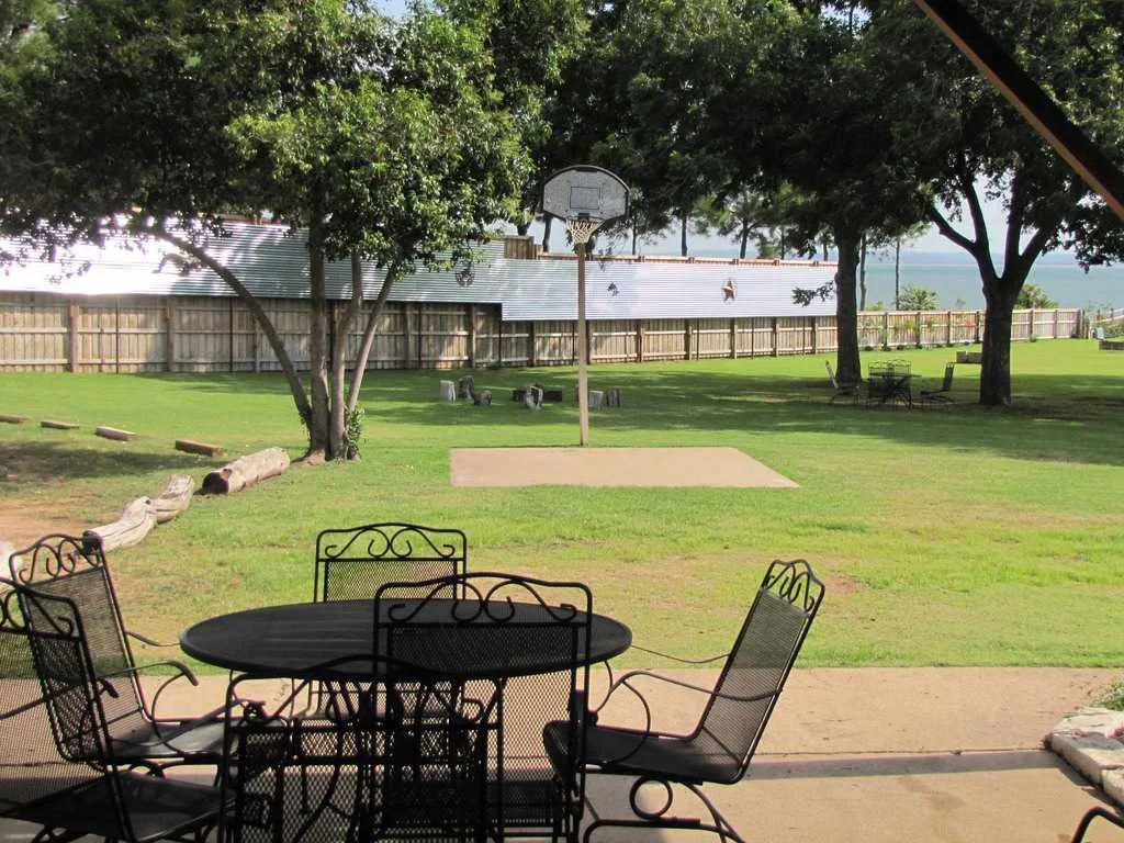 Backyard patio with black metal table and chairs, overlooking a grassy yard with a basketball hoop and a wooden fence. Trees provide shade and a lake is visible in the distance. Hill Country Texas lake cabin rentals.