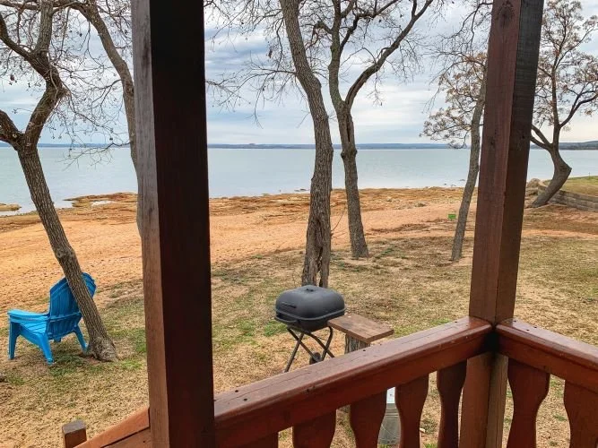 Lake view from a wooden porch with trees, a barbecue grill, and blue chairs on a sandy shore. Hill Country Texas lake cabin rentals.