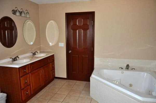 Bathroom with double sink vanity, two oval mirrors, a wooden door, and a white corner bathtub on tiled floor. Hill Country Texas lake cabin rentals.