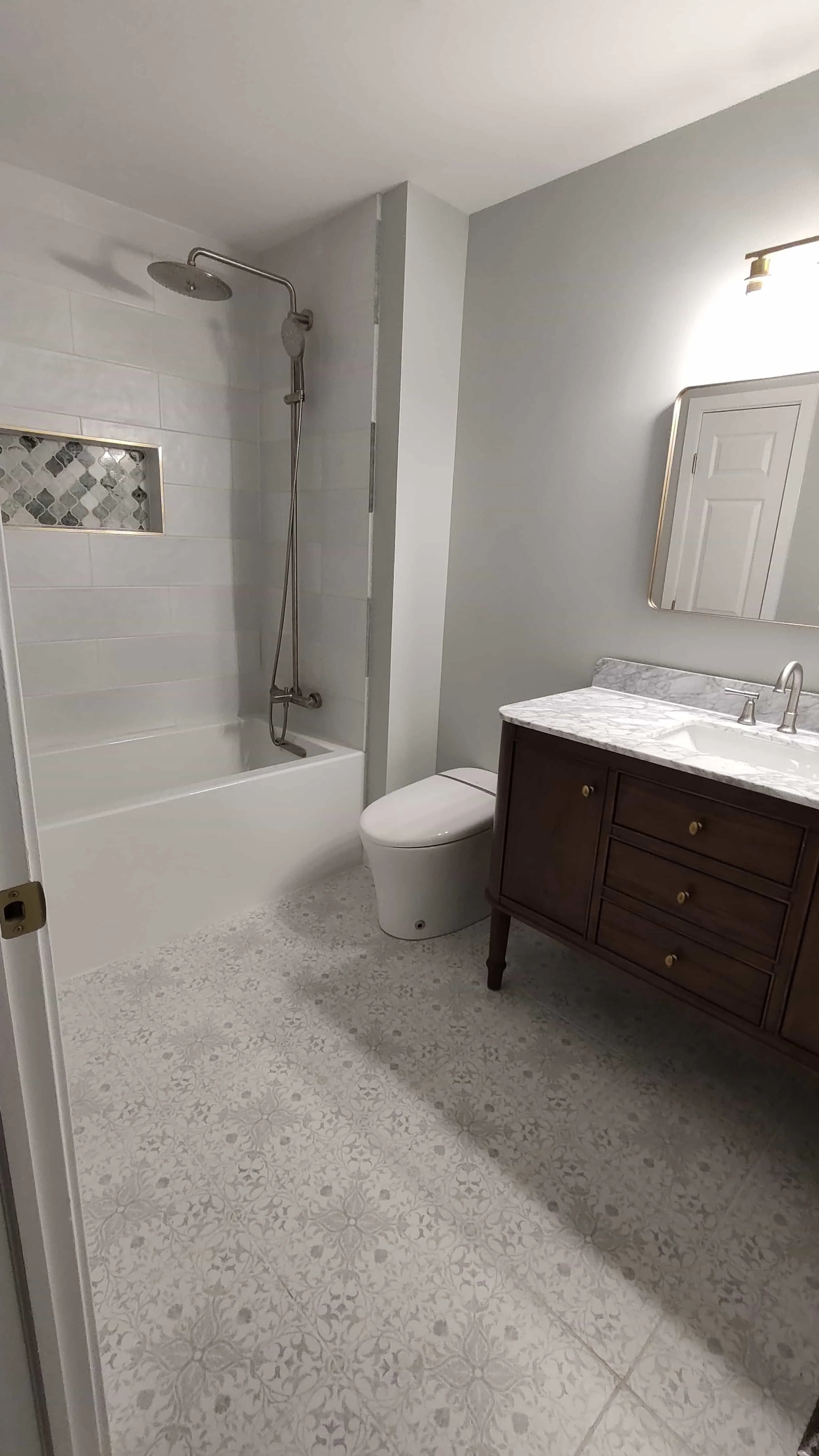 A modern bathroom featuring a bathtub with a shower, a toilet, and a wooden vanity with a marble countertop and an oval mirror, illuminated by a wall-mounted light.