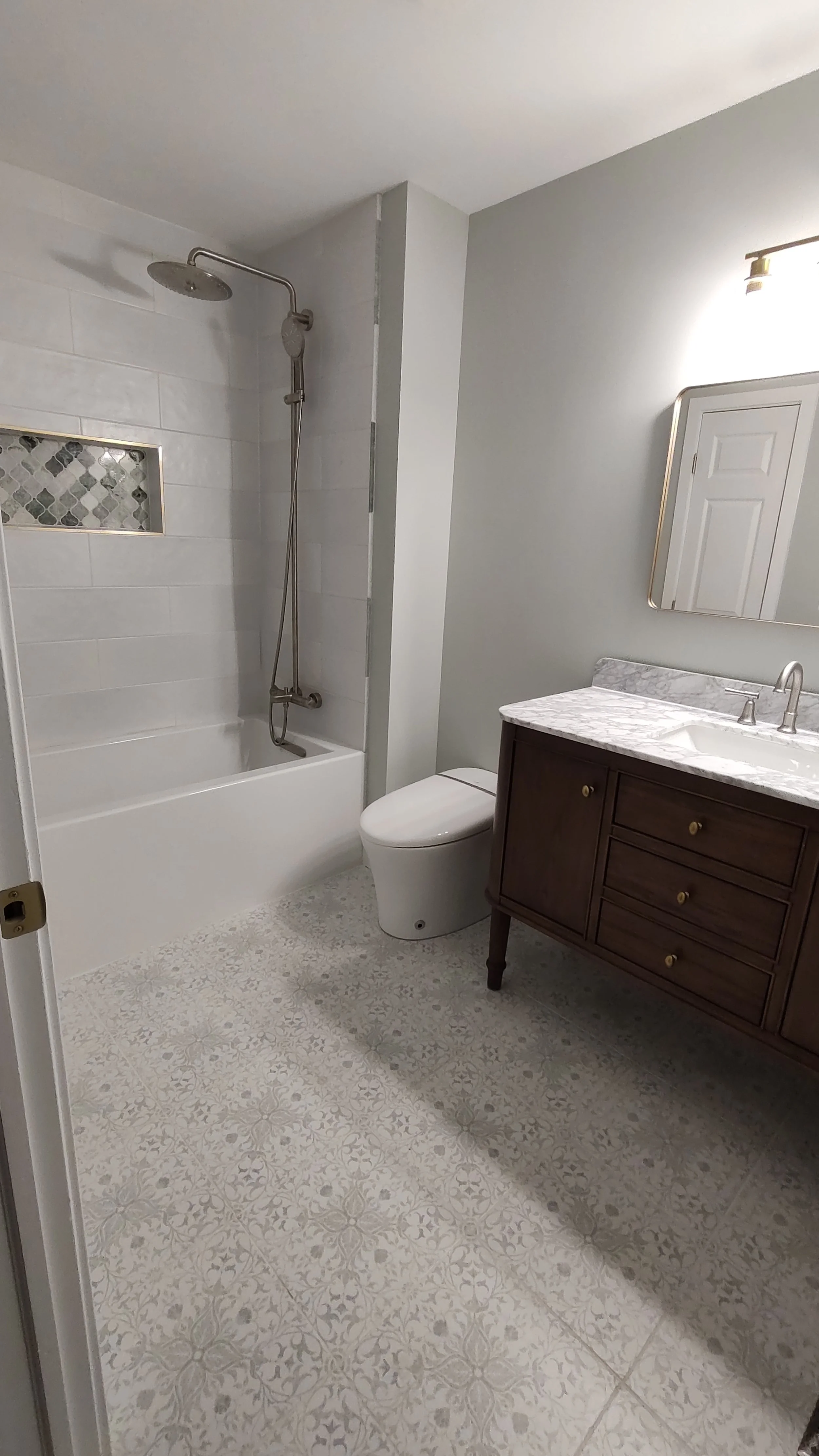 A modern bathroom featuring a bathtub with a shower, a toilet, and a wooden vanity with a marble countertop and an oval mirror, illuminated by a wall-mounted light.