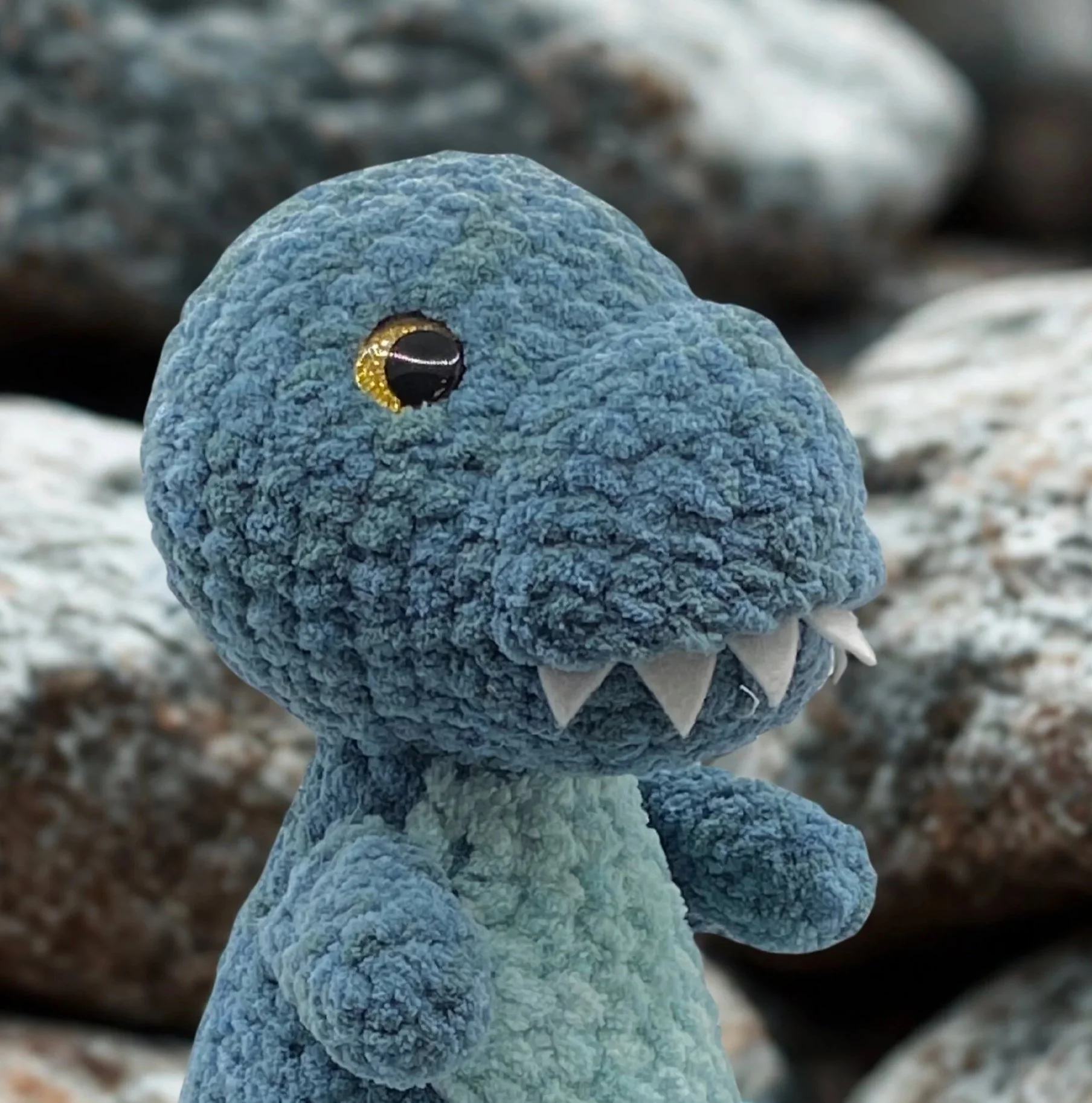 A hand crocheted plushie of a blue dinosaur, a large head, small arms, a rounded snout, a large black and brown eye, and small white teeth, positioned in front of a background of rocks.