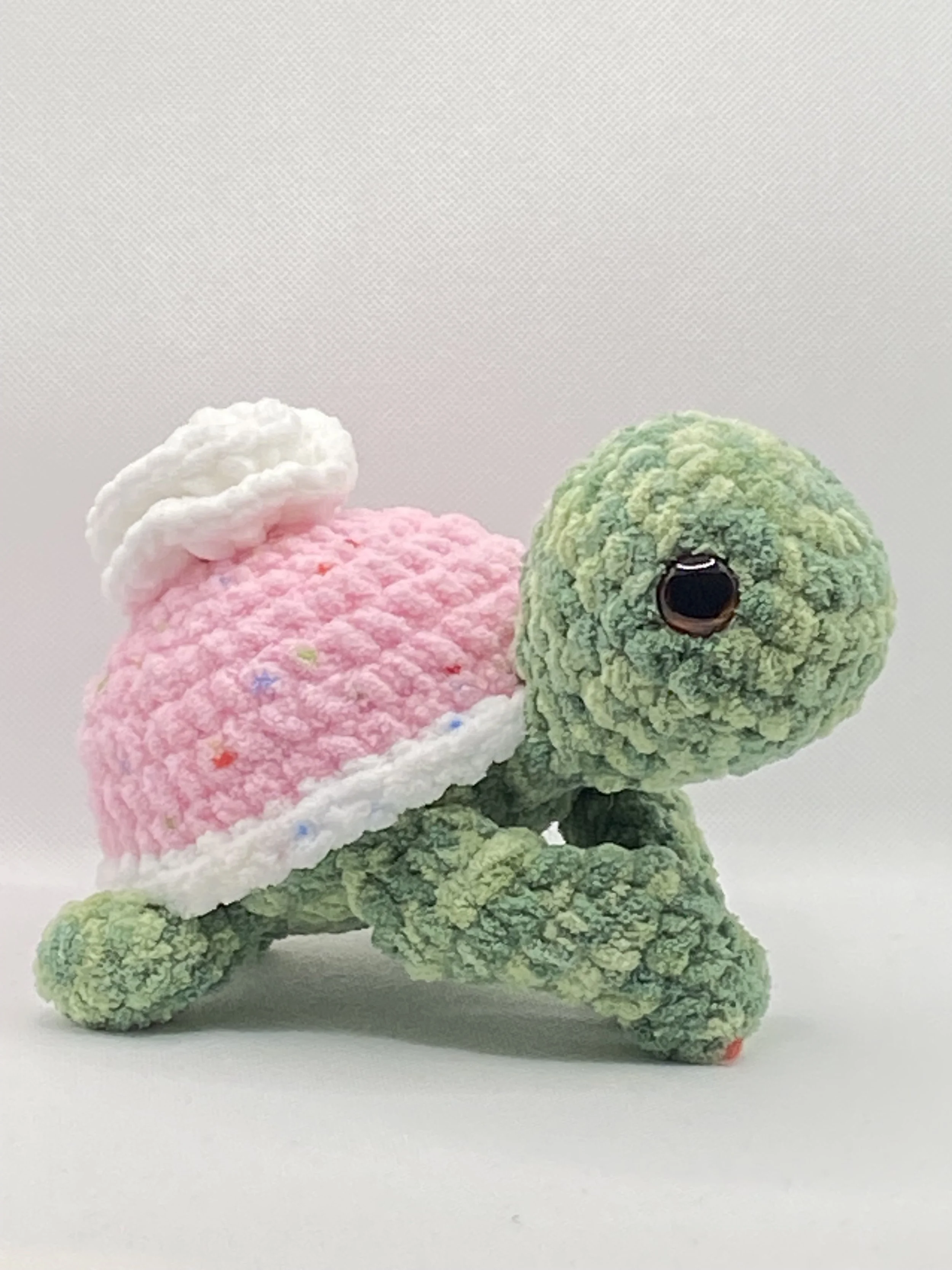 A plush crocheted green turtle with a pink shell and a white trim, set against a plain background.