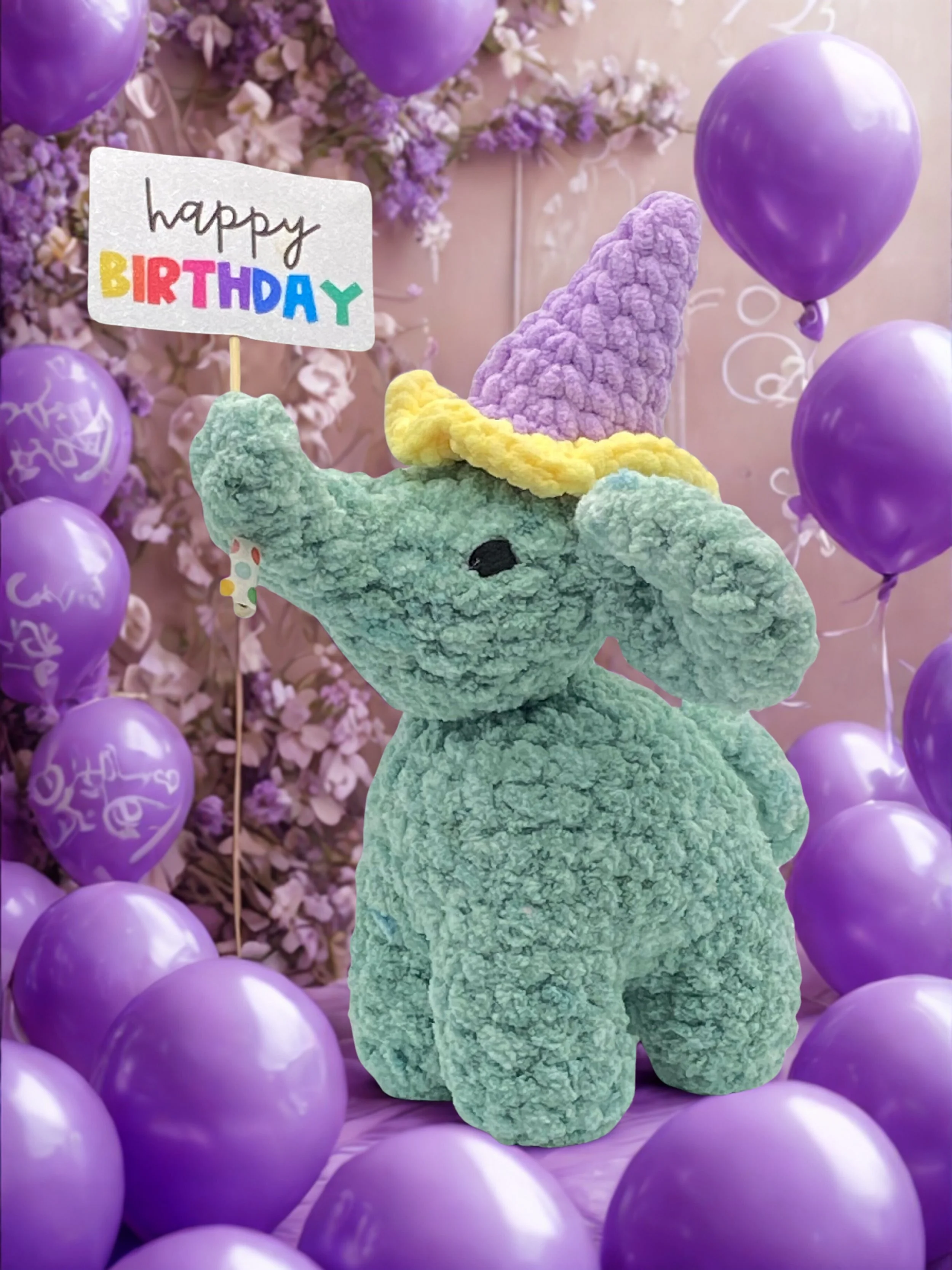 A plush elephant toy with a pastel purple, yellow, and blue hat , surrounded by purple balloons and pink flower decorations, holding a handmade 'happy birthday' sign.