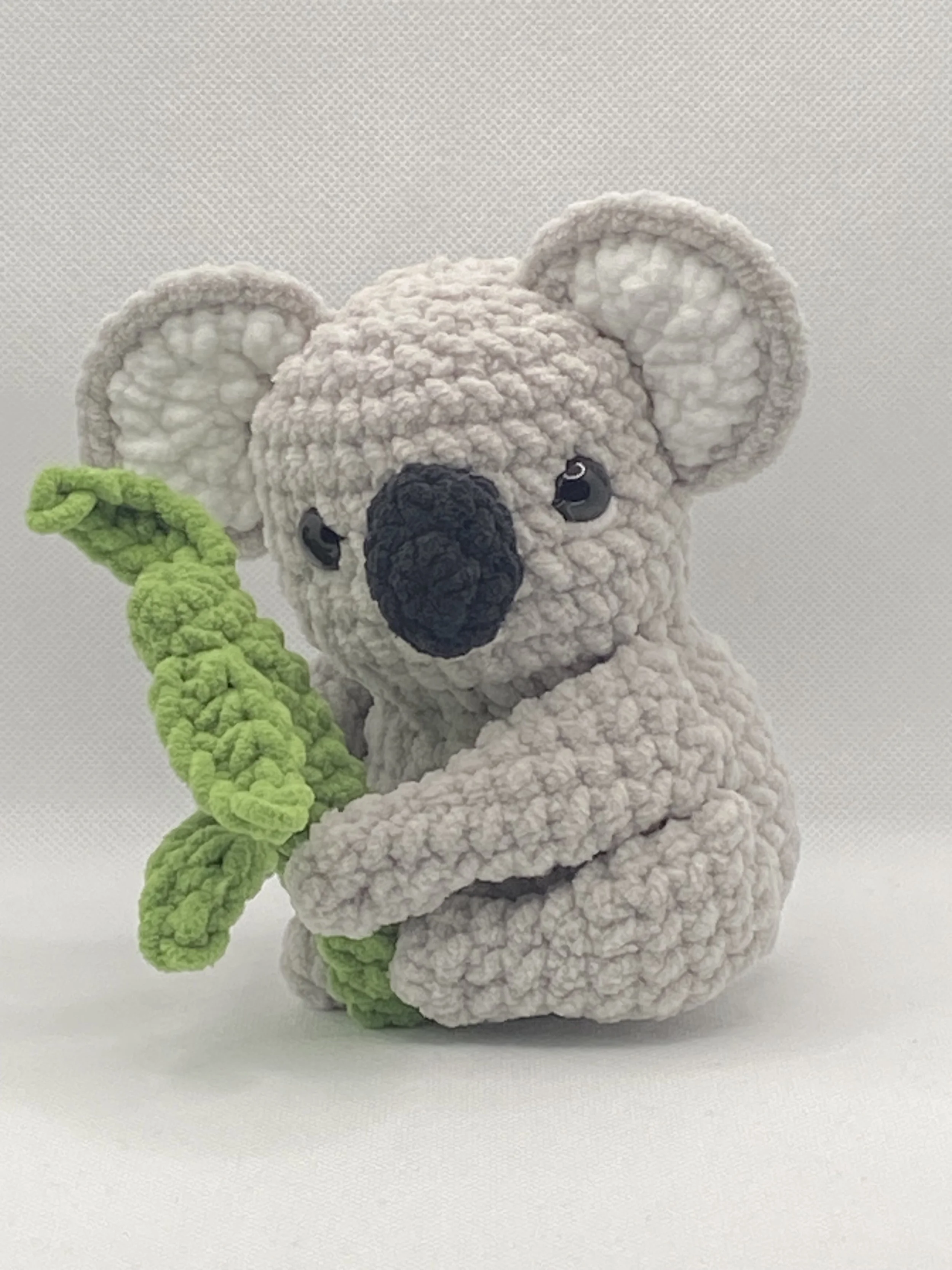 Crocheted koala holding a green leaf, with a white textured background.