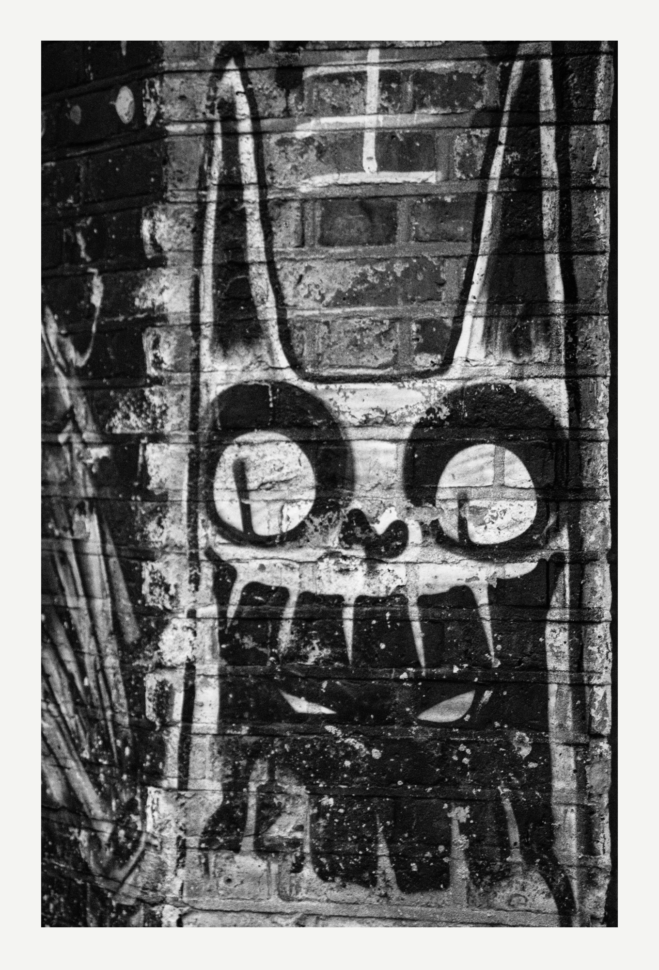 Black and white graffiti art on a brick wall depicting a stylized face with large eyes, a nose, and a mouth, with visible spray paint drips and textures.