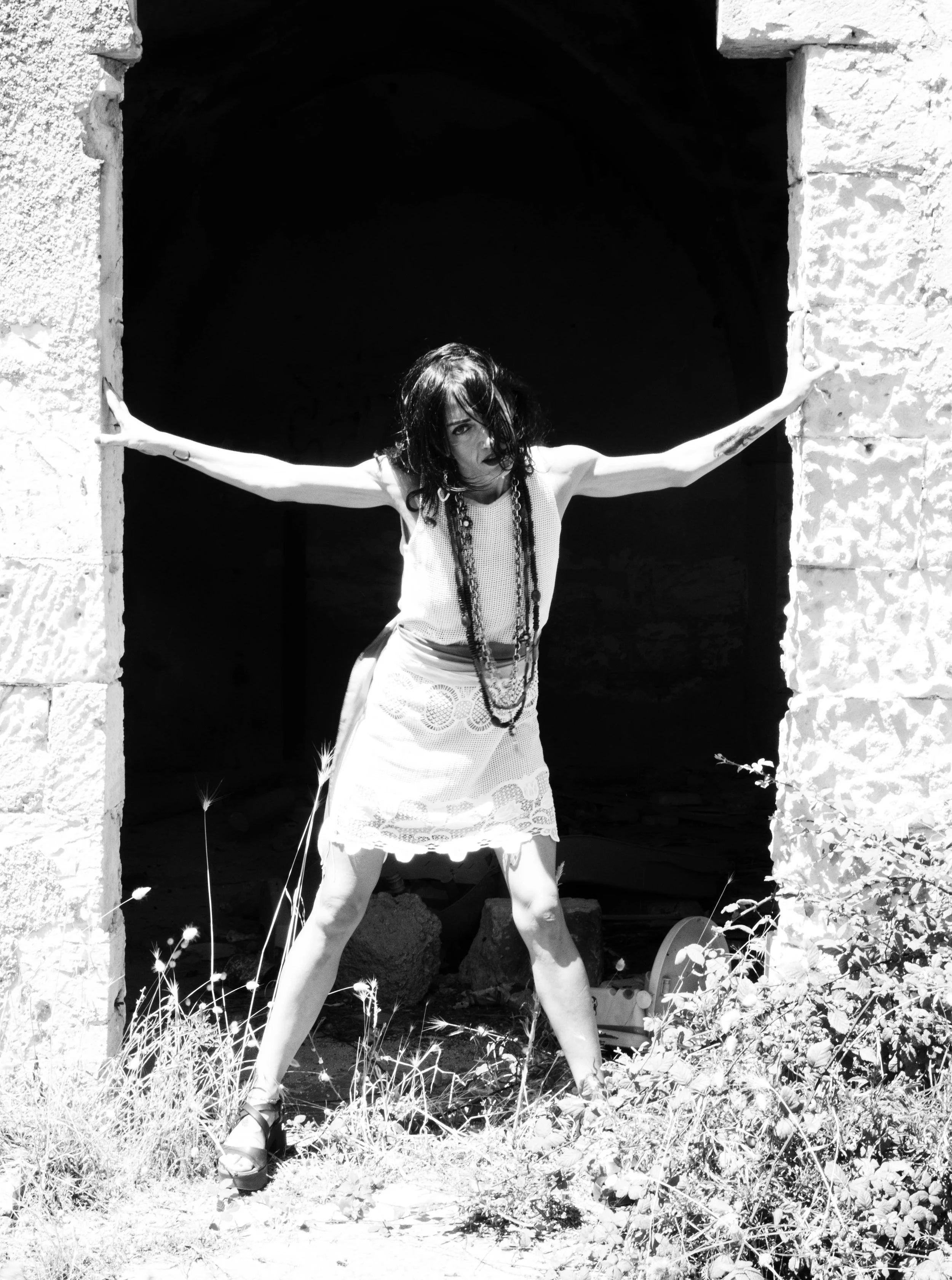 A woman with dark, wavy hair and tattoos is standing in front of an arched stone doorway with her arms outstretched and her hands touching the stone edges. She is wearing a sleeveless top, a skirt, and high-heeled shoes, with necklaces around her neck. The photo is in black and white, and the woman is standing outdoors, surrounded by wild plants and grasses.