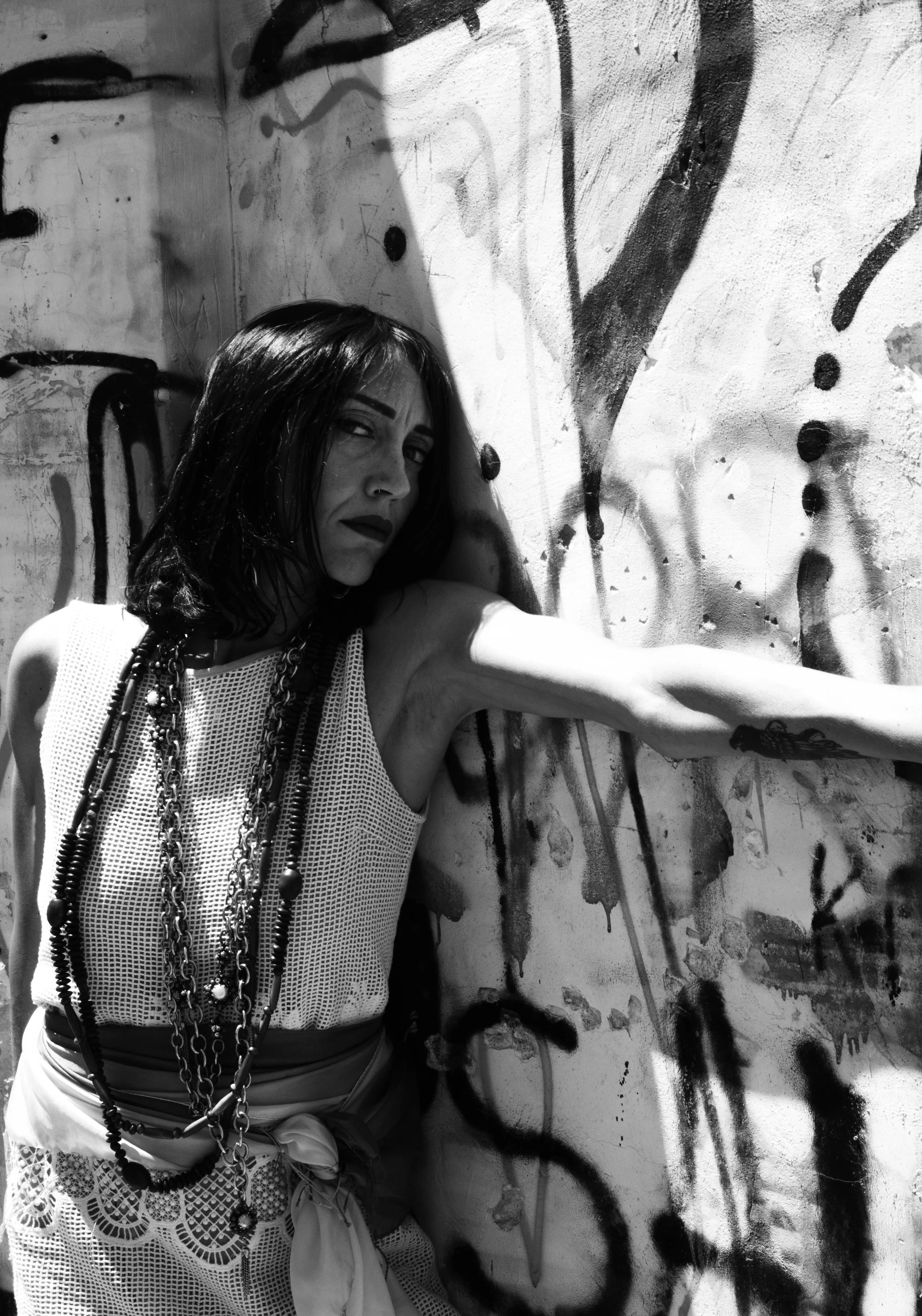 A woman with dark hair and makeup, wearing a sleeveless white mesh top and multiple necklaces, takes a selfie against a graffiti-covered wall with drips and splatters in black and white.