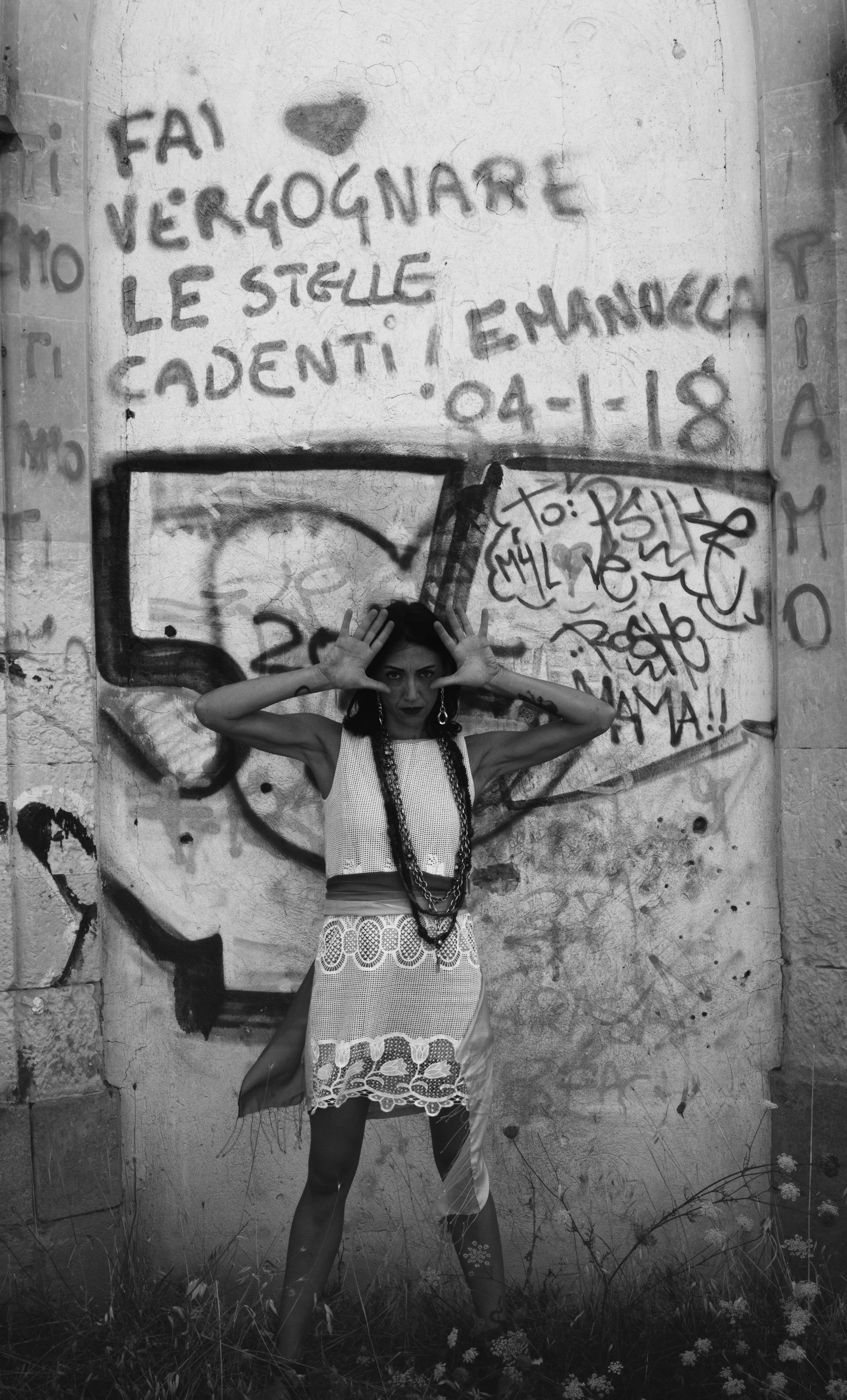A woman standing in front of a graffiti-covered wall. She has short dark hair, wears a sleeveless lace dress, and accessorizes with multiple necklaces and earrings. Her hands frame her face.