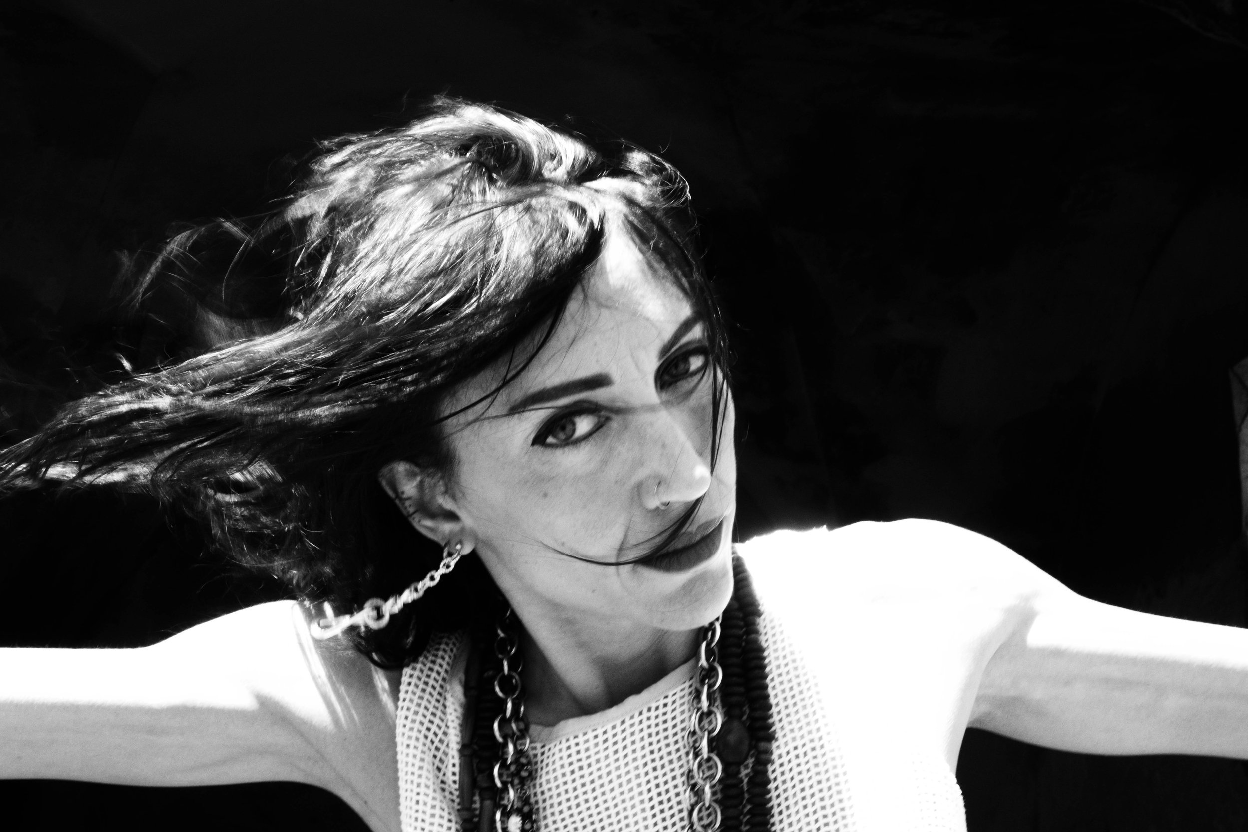 Black and white photo of a young woman with curly hair, looking at the camera, wearing a chain necklace and earrings, with dark background.