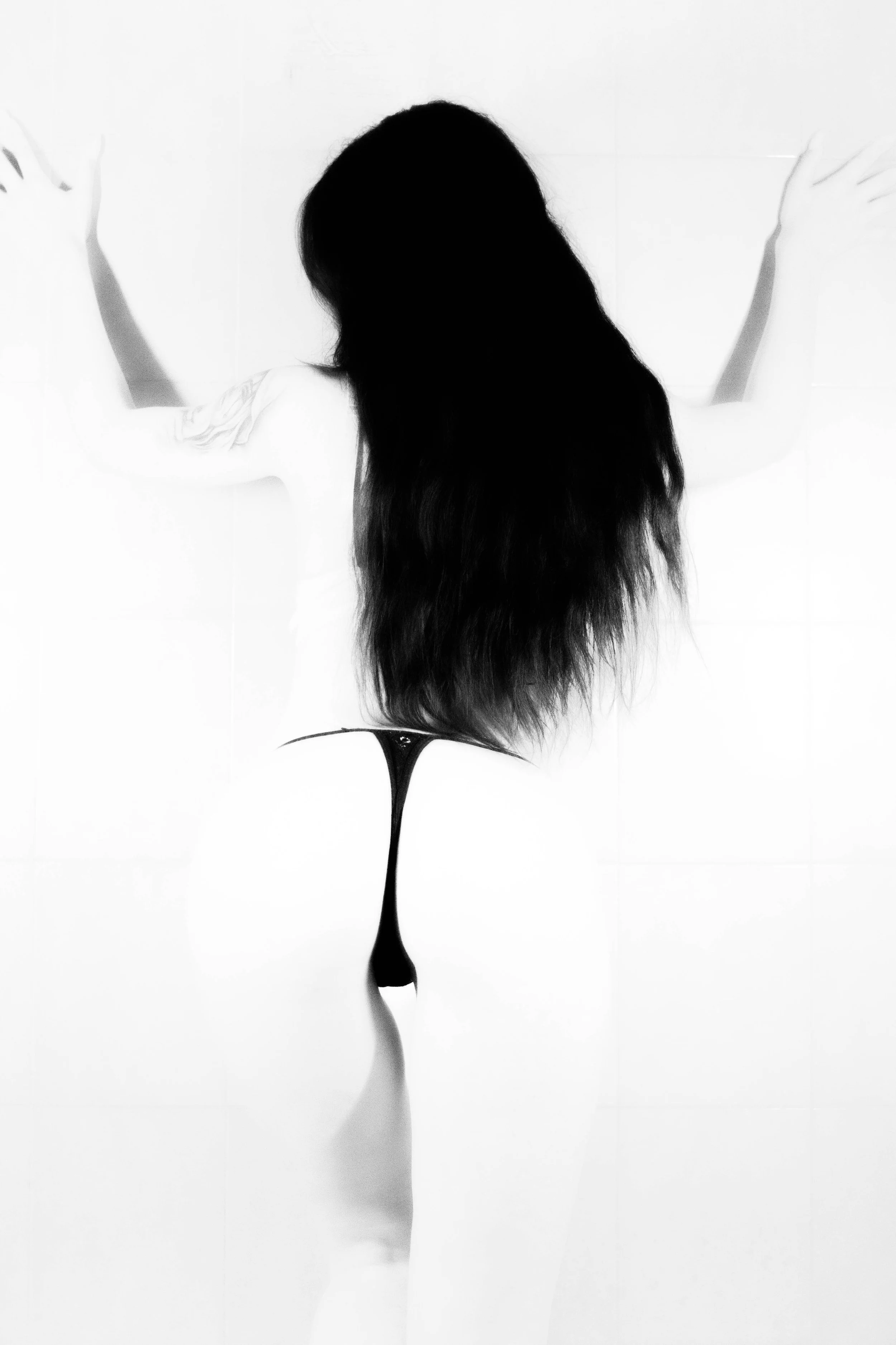 Black and white photo of a woman with long hair facing away, wearing a thong, with her arms raised and hands touching the wall.