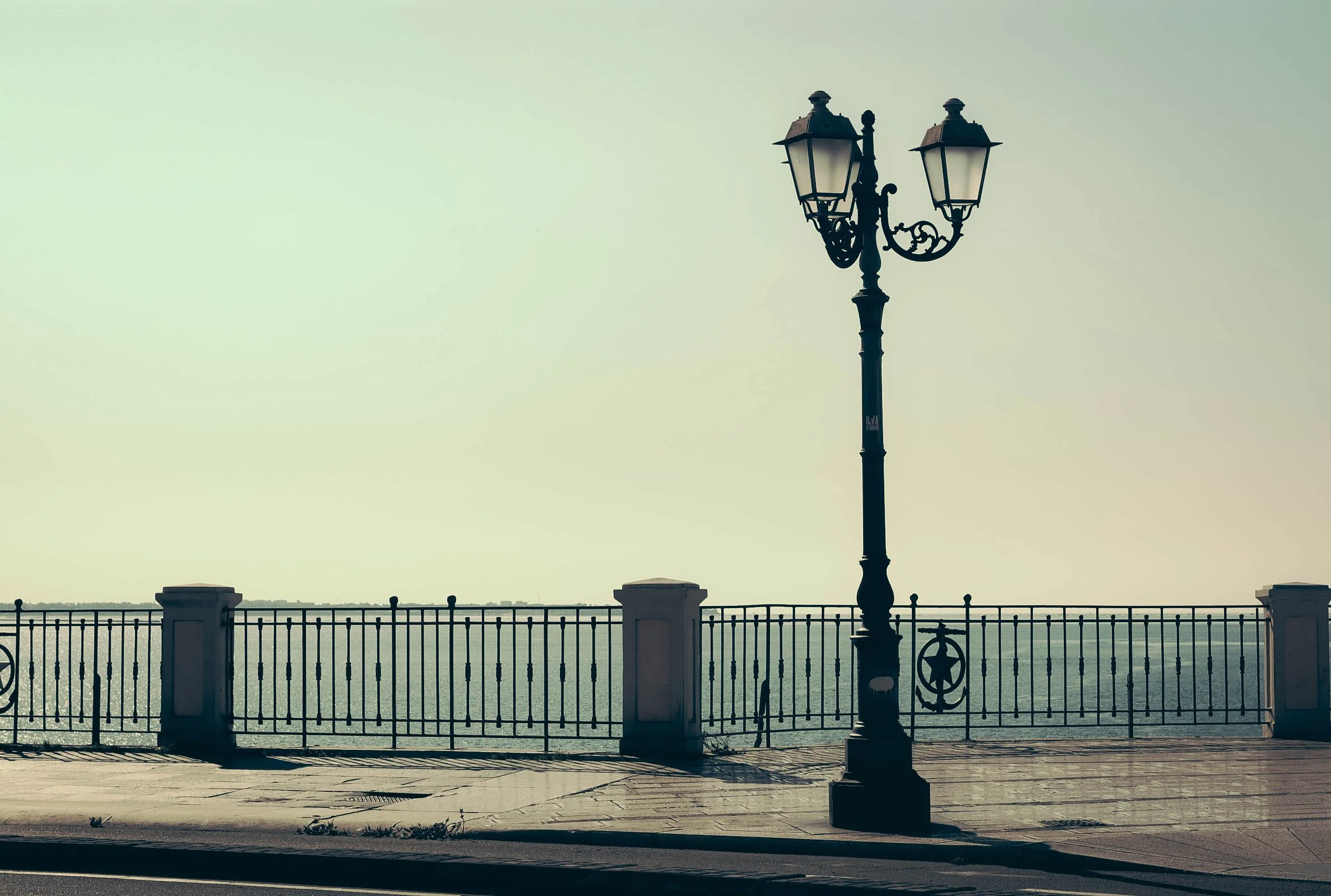 A vintage street lamp with two lanterns, positioned along a waterfront promenade with a metal fence in the foreground. The scene is set against a hazy sky over a body of water.
