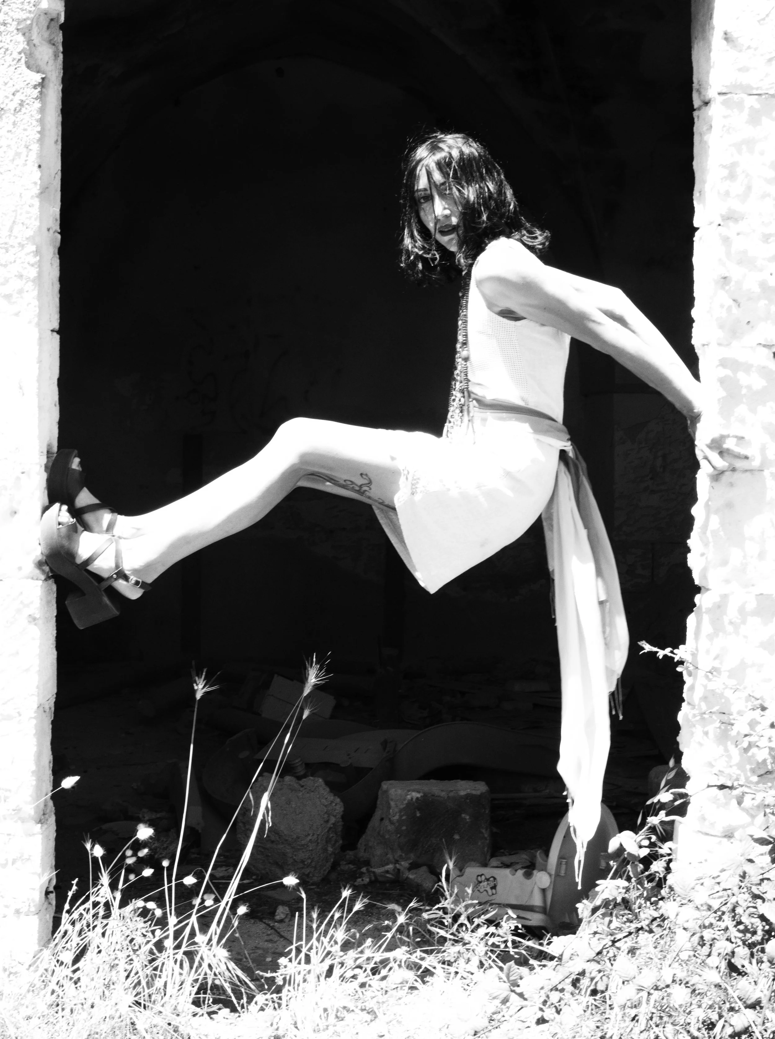 Black and white photo of a woman with dark, shoulder-length hair, wearing a sleeveless top and a long skirt, sitting on a ledge in a dark, empty doorway. She has one leg extended and the other bent, with her foot resting on the ledge. The woman is looking towards the camera with an intense expression. There are plants and flowers in the foreground.