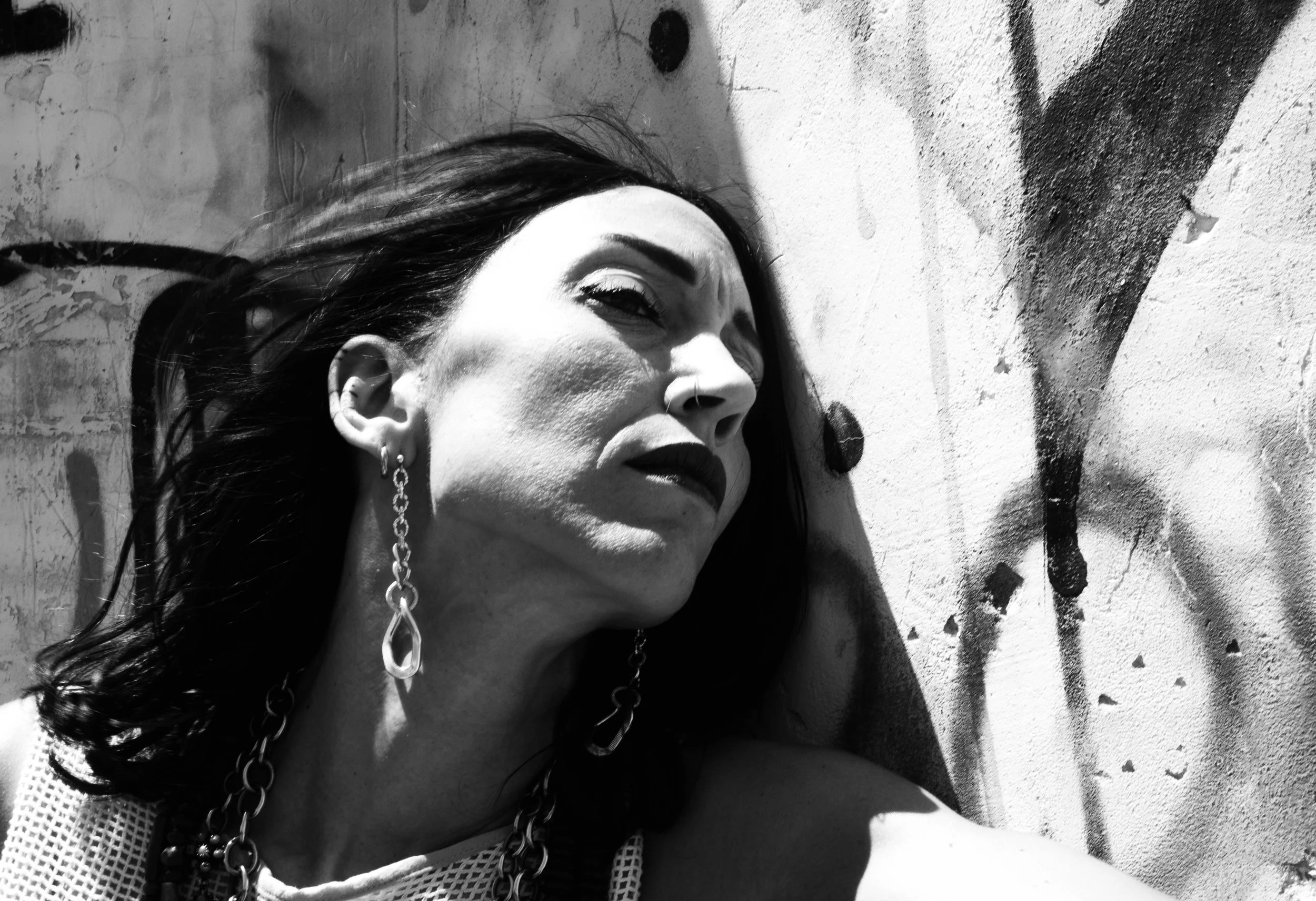 A woman with dark hair and earrings leaning against a graffiti-covered wall in black and white