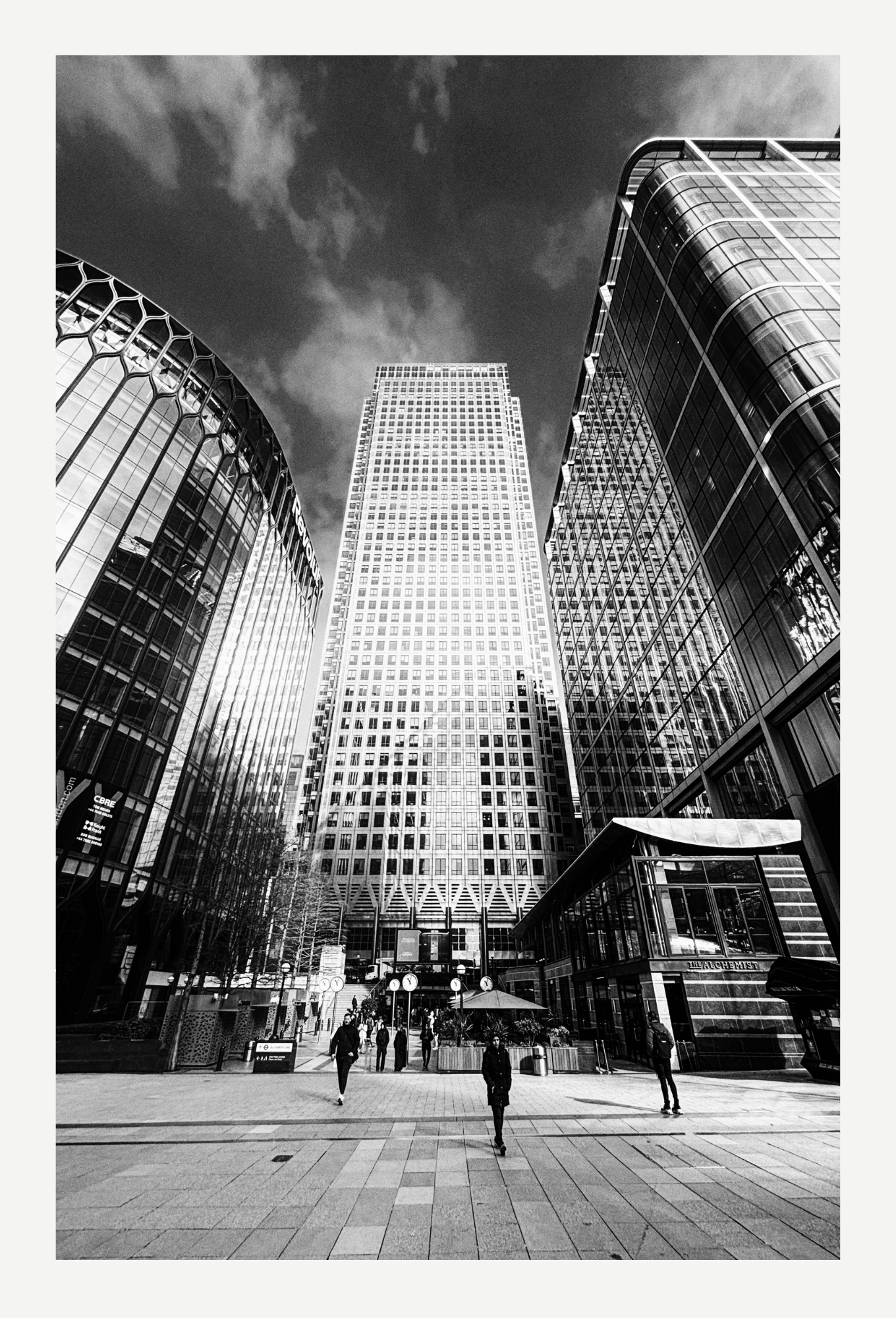 Cityscape with tall modern buildings, people walking in a plaza, some trees, and outdoor seating, black and white photo.