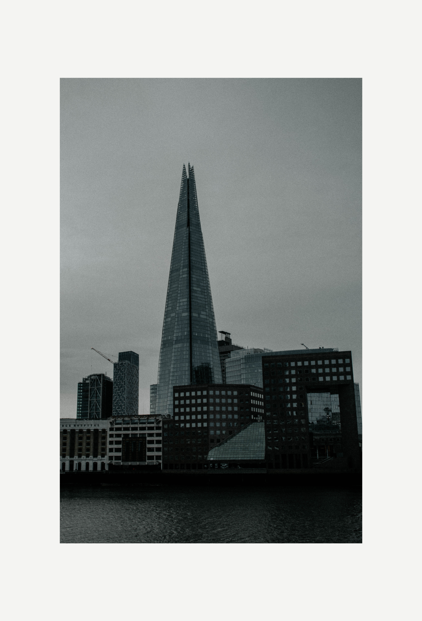 A tall, modern skyscraper with a pointed top in a cityscape, surrounded by other high-rise buildings, with water in the foreground and a cloudy sky overhead.