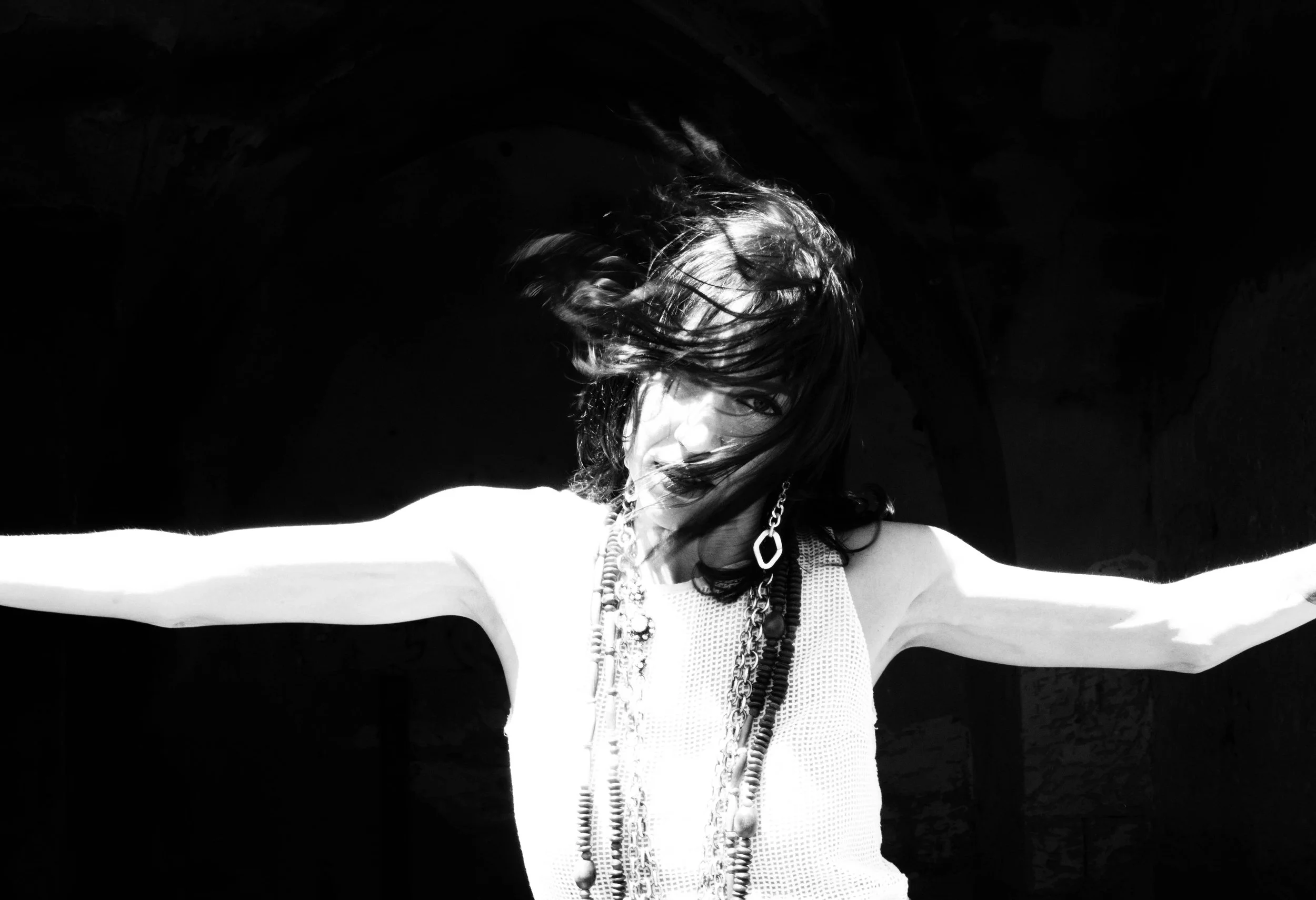 A woman with long hair wearing a necklace and a sleeveless top stretching her arms out, with her hair covering part of her face, against a dark background.