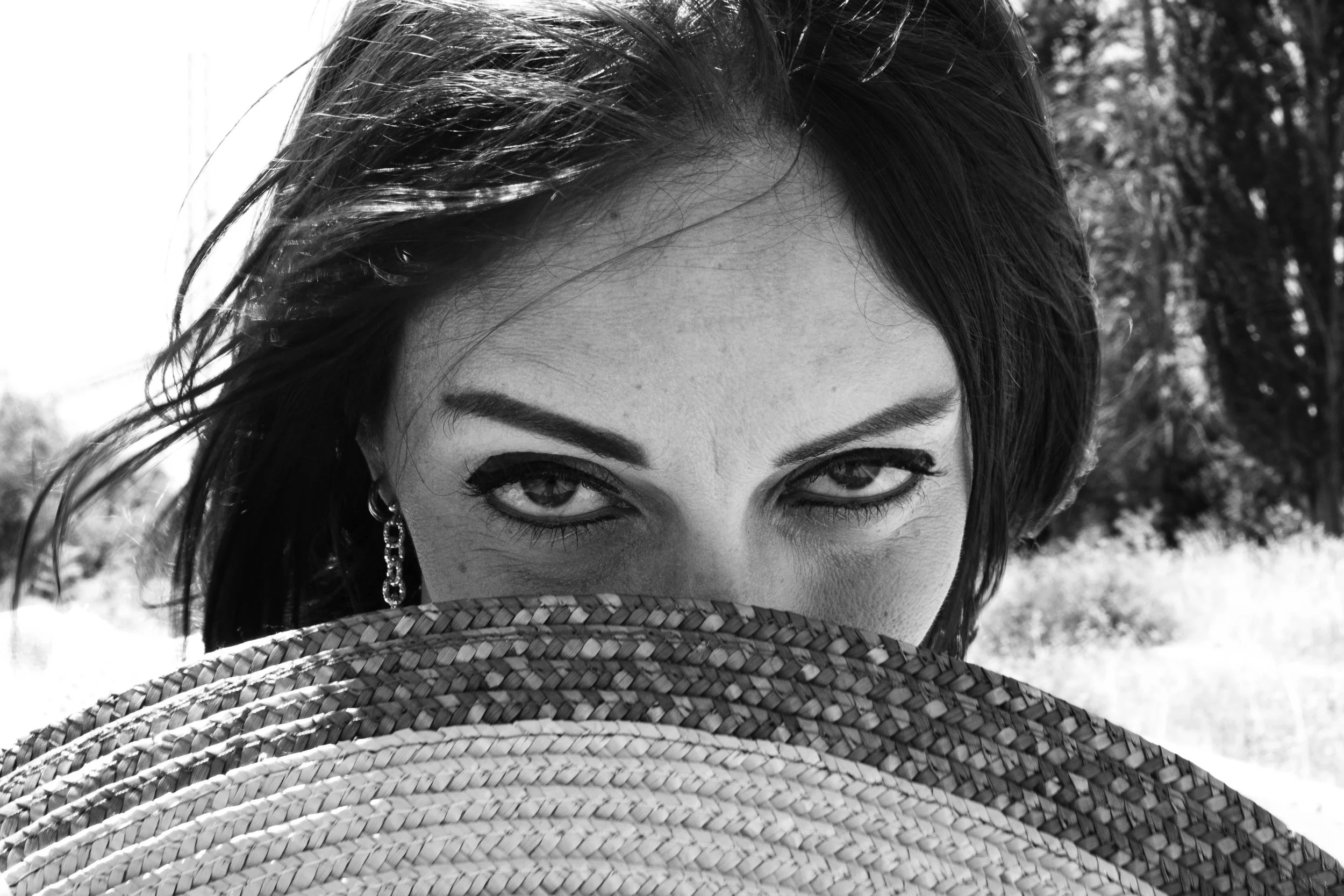 A black and white photo of a person with dark hair, expressive eyes, wearing earrings, partially covered by a woven hat and face covering, outdoors with trees in the background.