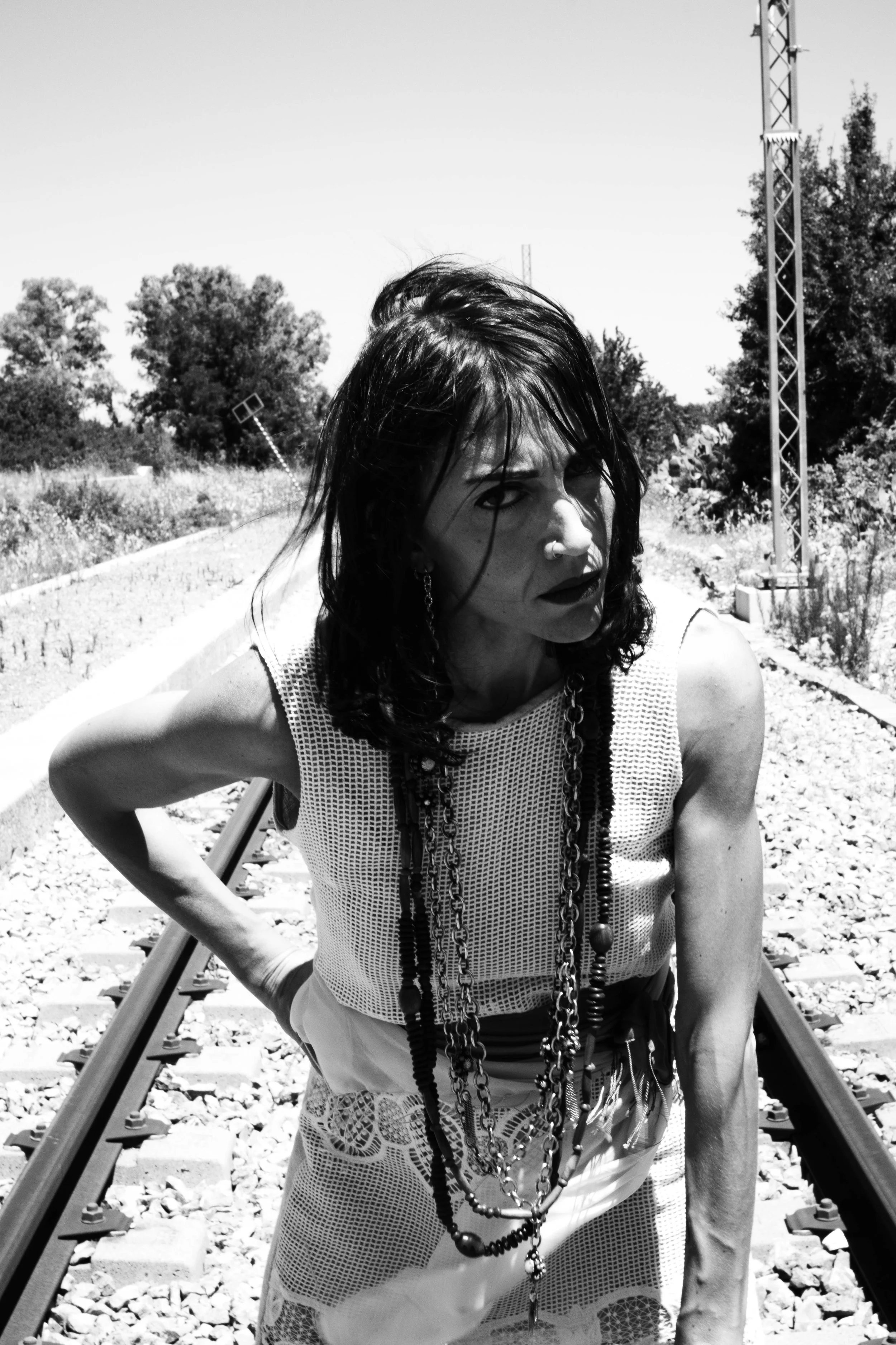 A woman with dark, wet hair and multiple necklaces, wearing a sleeveless, patterned top, standing on a railroad track outdoors with trees and a metal structure in the background.