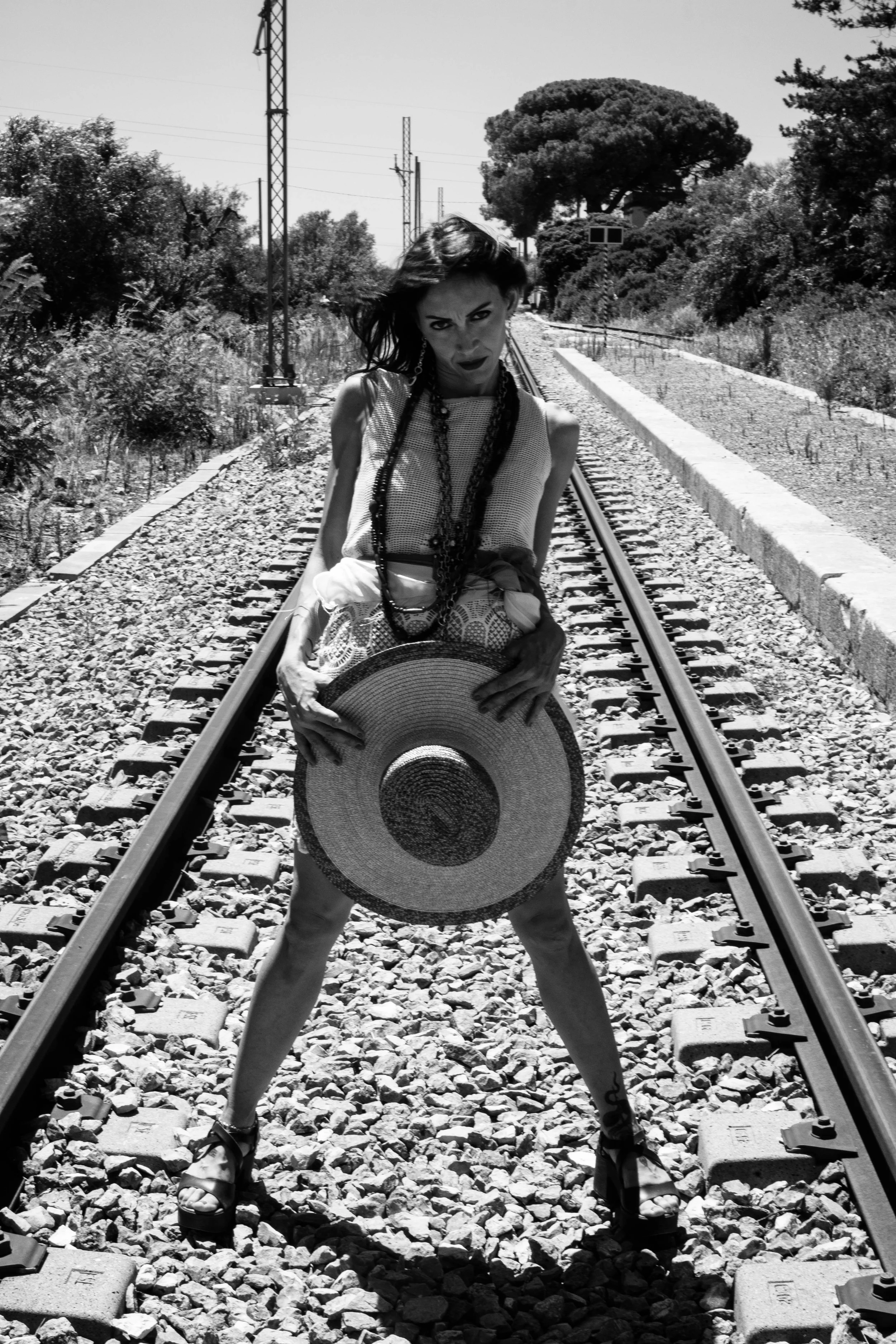 A woman standing on railway tracks holding a large straw hat, with trees and power lines in the background, in black and white.