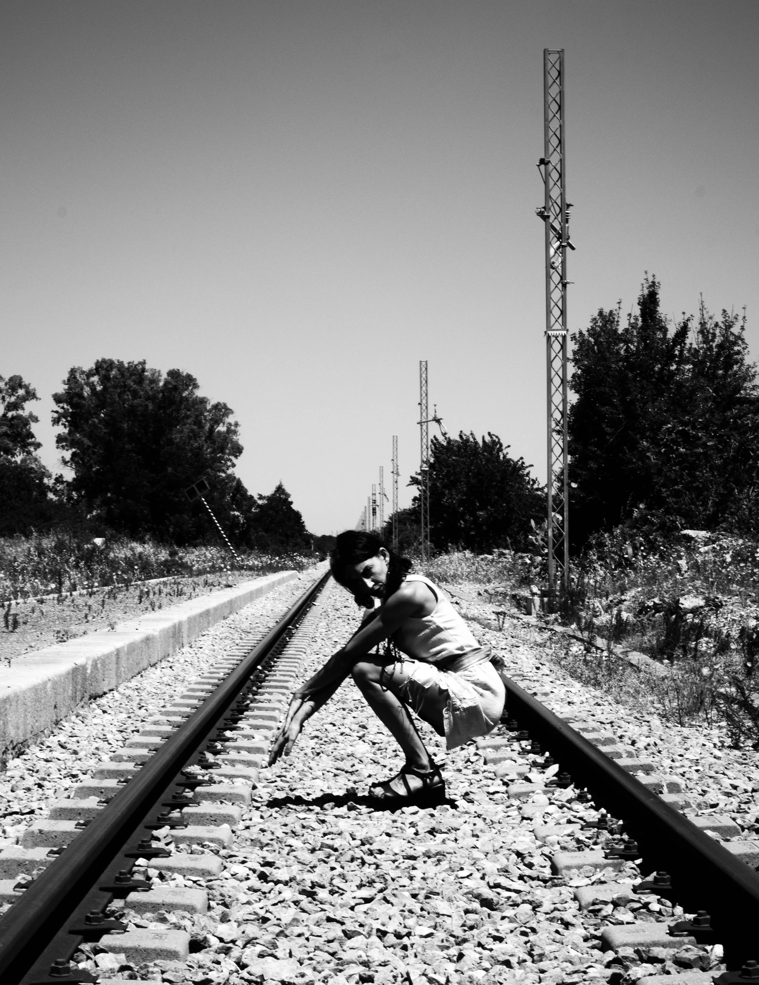 A woman in casual clothing and sandals crouches on a railway track among trees and telephone poles, with her head tilted downward and her arms resting on her knees.