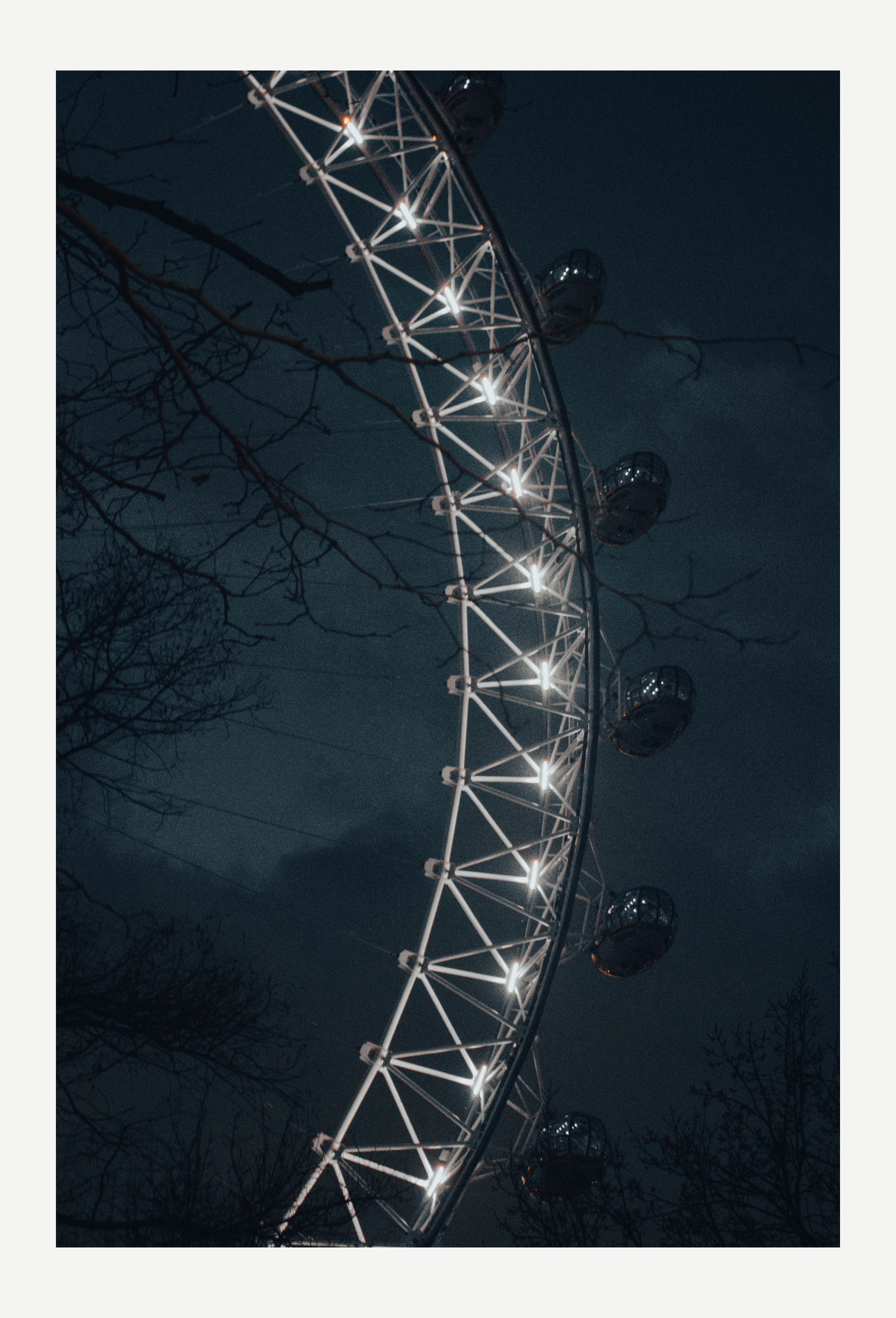 Nighttime view of a Ferris wheel illuminated with bright lights, with leafless tree branches silhouetted in the foreground against a dark sky.