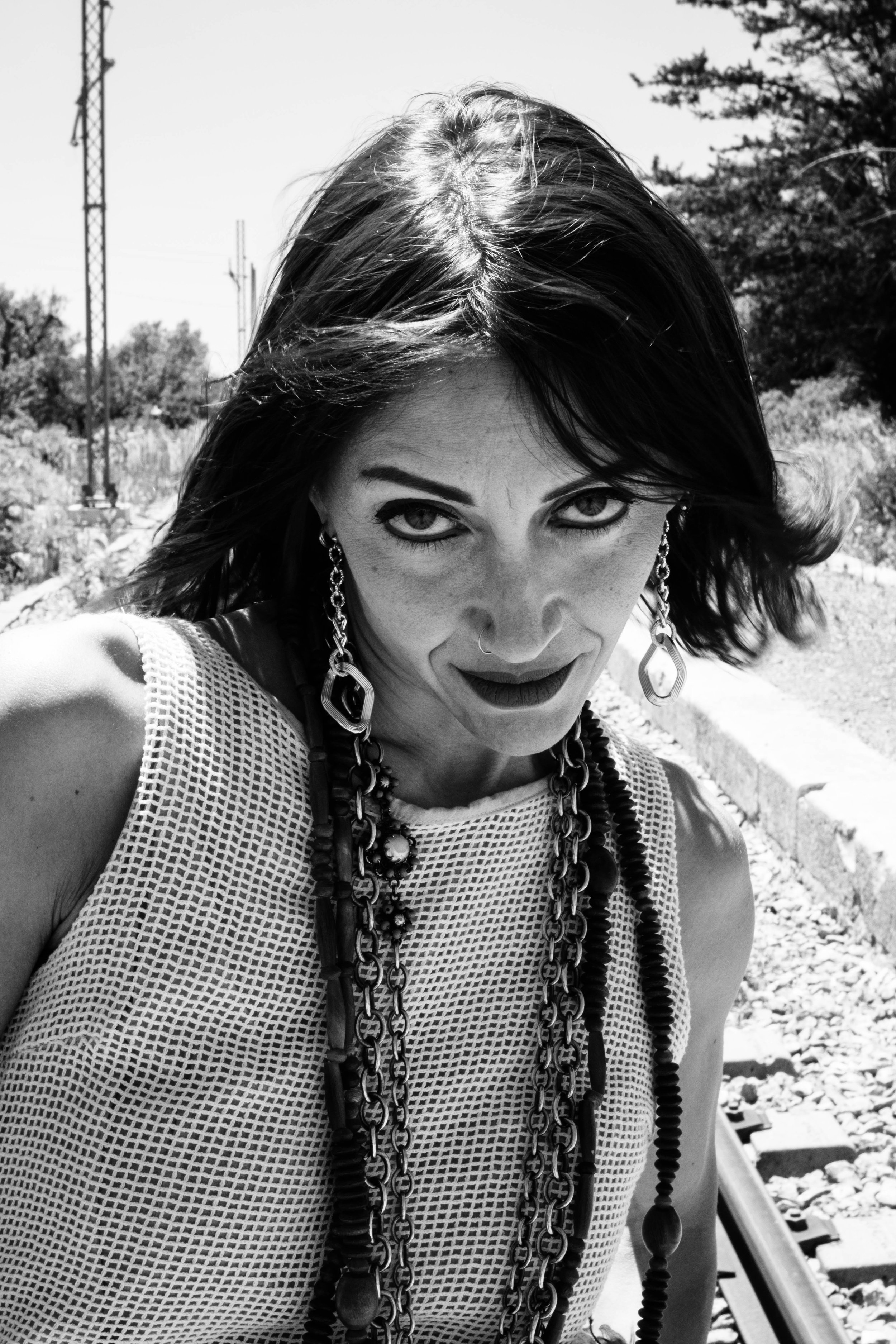 A black and white photo of a woman with shoulder-length hair, wearing large earrings, layered necklaces, and a sleeveless, netted top. She is outdoors near train tracks, with trees and power lines in the background, looking directly at the camera.