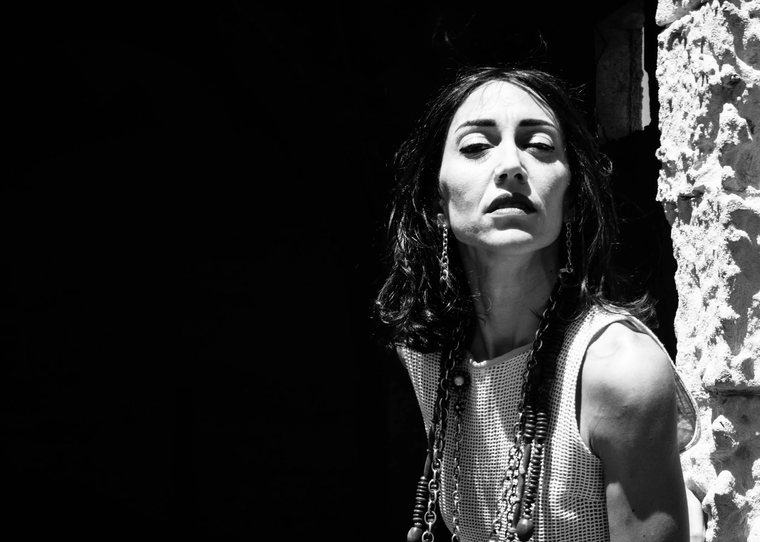 A woman with dark, wavy hair and earrings posing outdoors against a stone wall, with strong sunlight highlighting her face and sleeveless top.