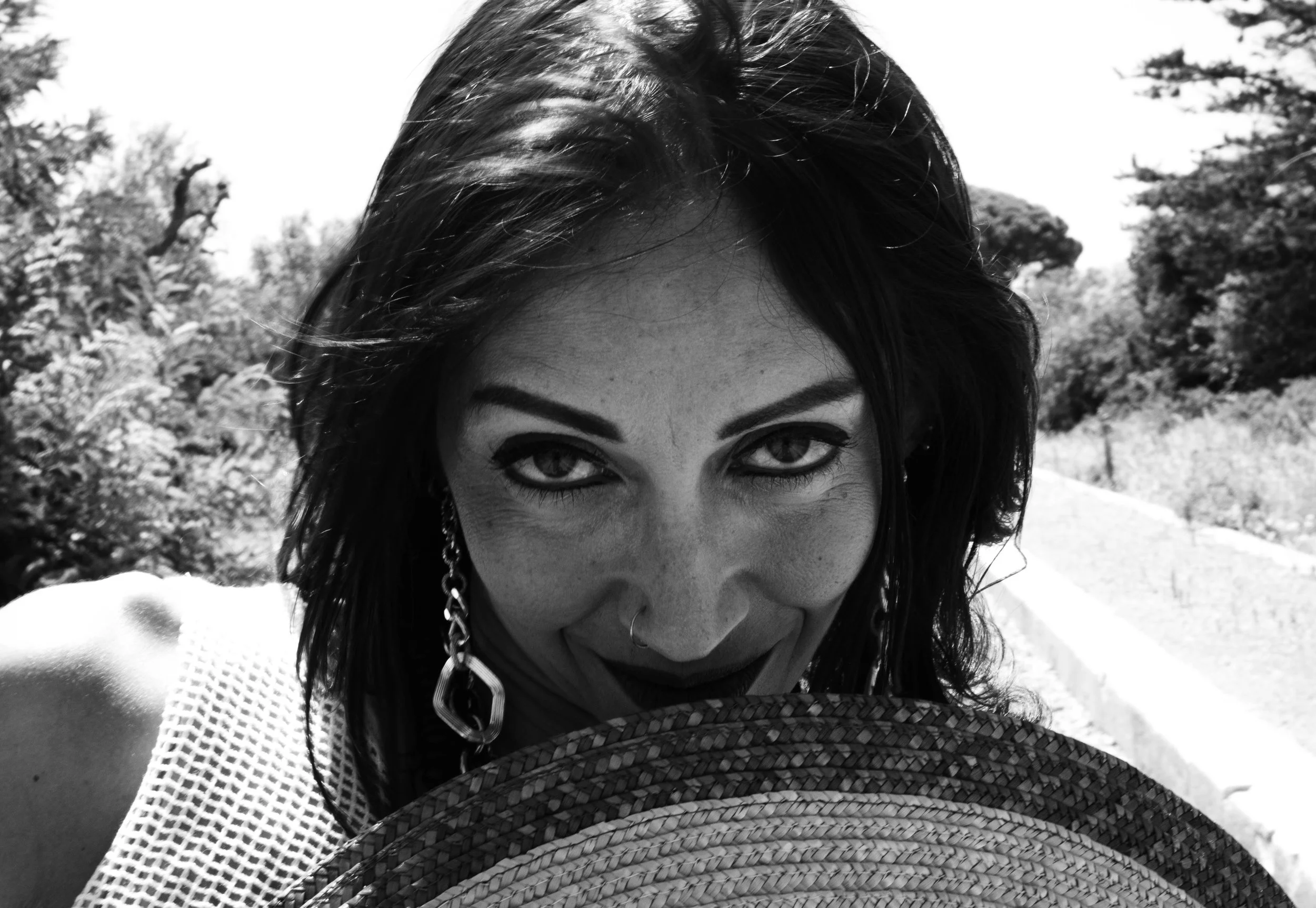 A black and white close-up photo of a smiling woman with dark hair, outdoors with trees in the background, wearing a hat and a white mesh top.