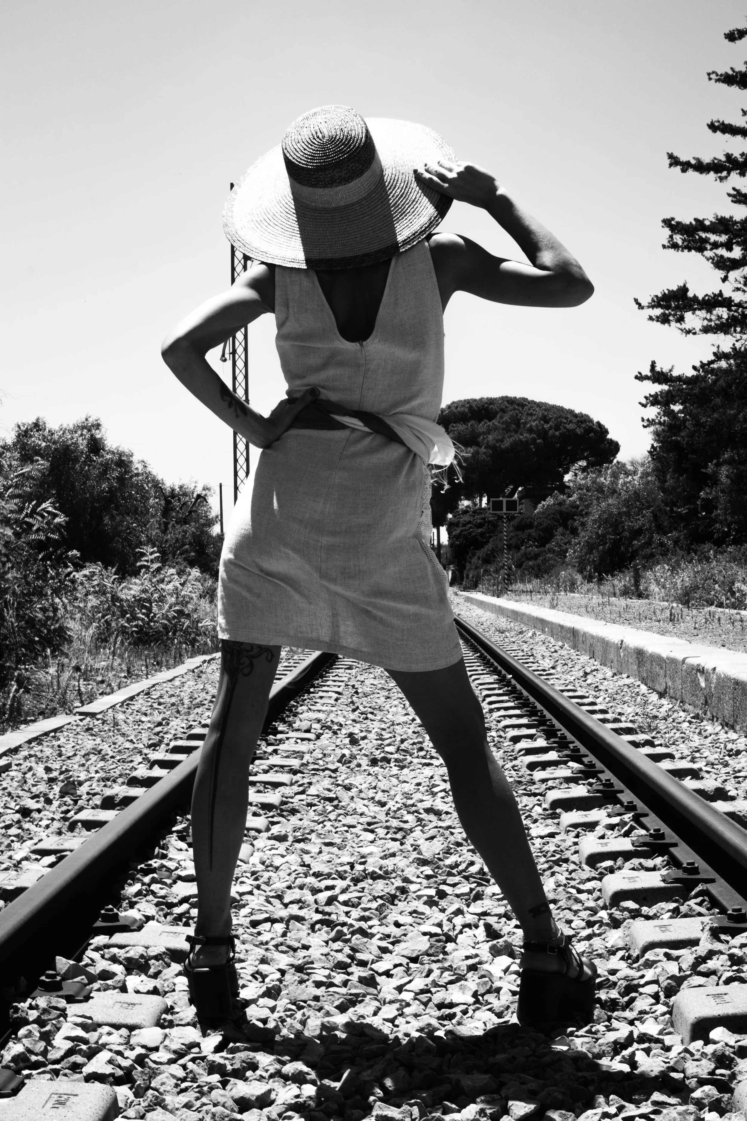 A person standing on train tracks, wearing a large sunhat and dress, with one hand on their hip and the other adjusting the hat, outdoors with trees in the background