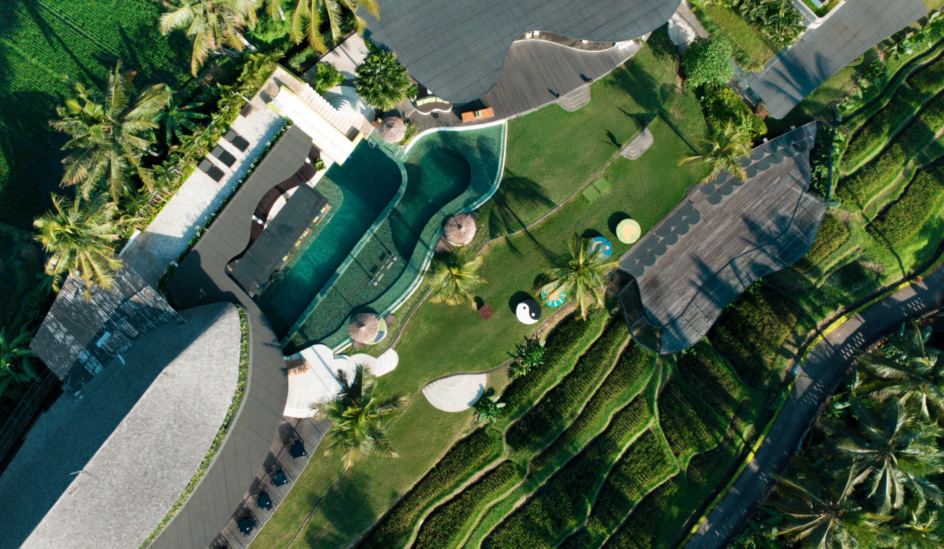 Bird's-eye view of a tropical resort with a large pool, surrounded by palm trees, greenery, and lounge chairs. There are pathways, thatched umbrellas, and circular decorative elements in the grass.