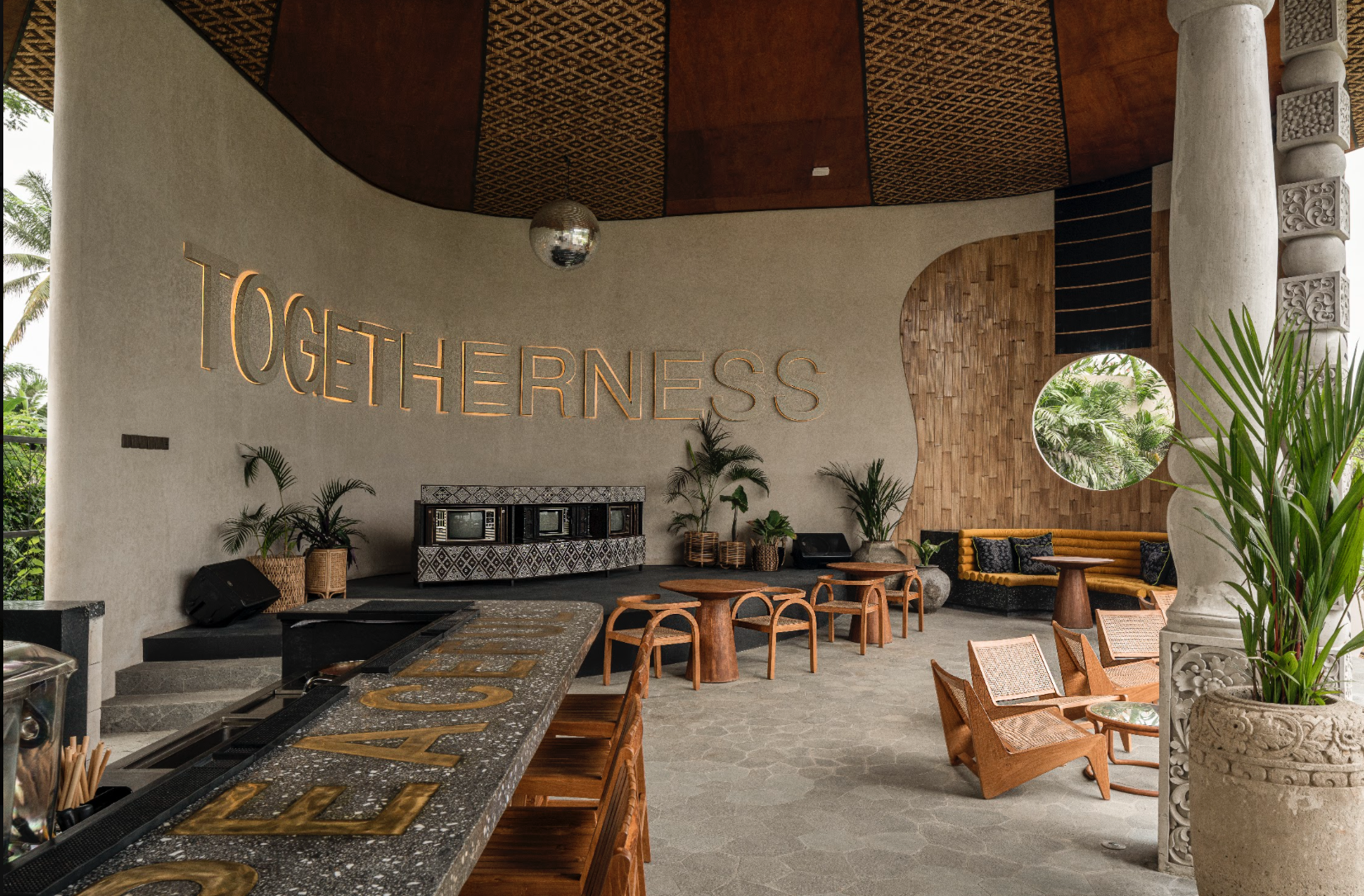 Interior of a tropical-themed lounge or bar area with the word 'TOGETHERNESS' illuminated on the wall, surrounded by potted plants, wooden furniture, a textured wall, and a circular window showing greenery outside.