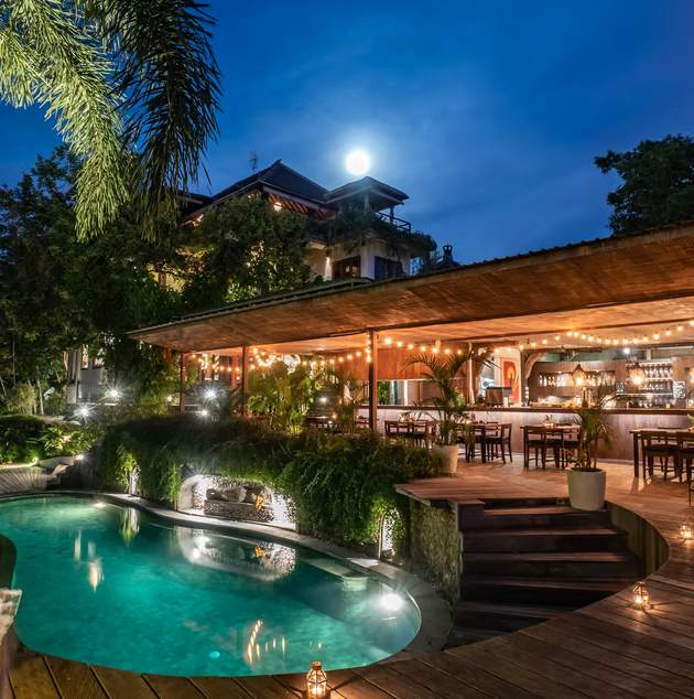 Night view of a luxurious outdoor restaurant beside a swimming pool, with warm string lights and lush greenery, under a moonlit sky.