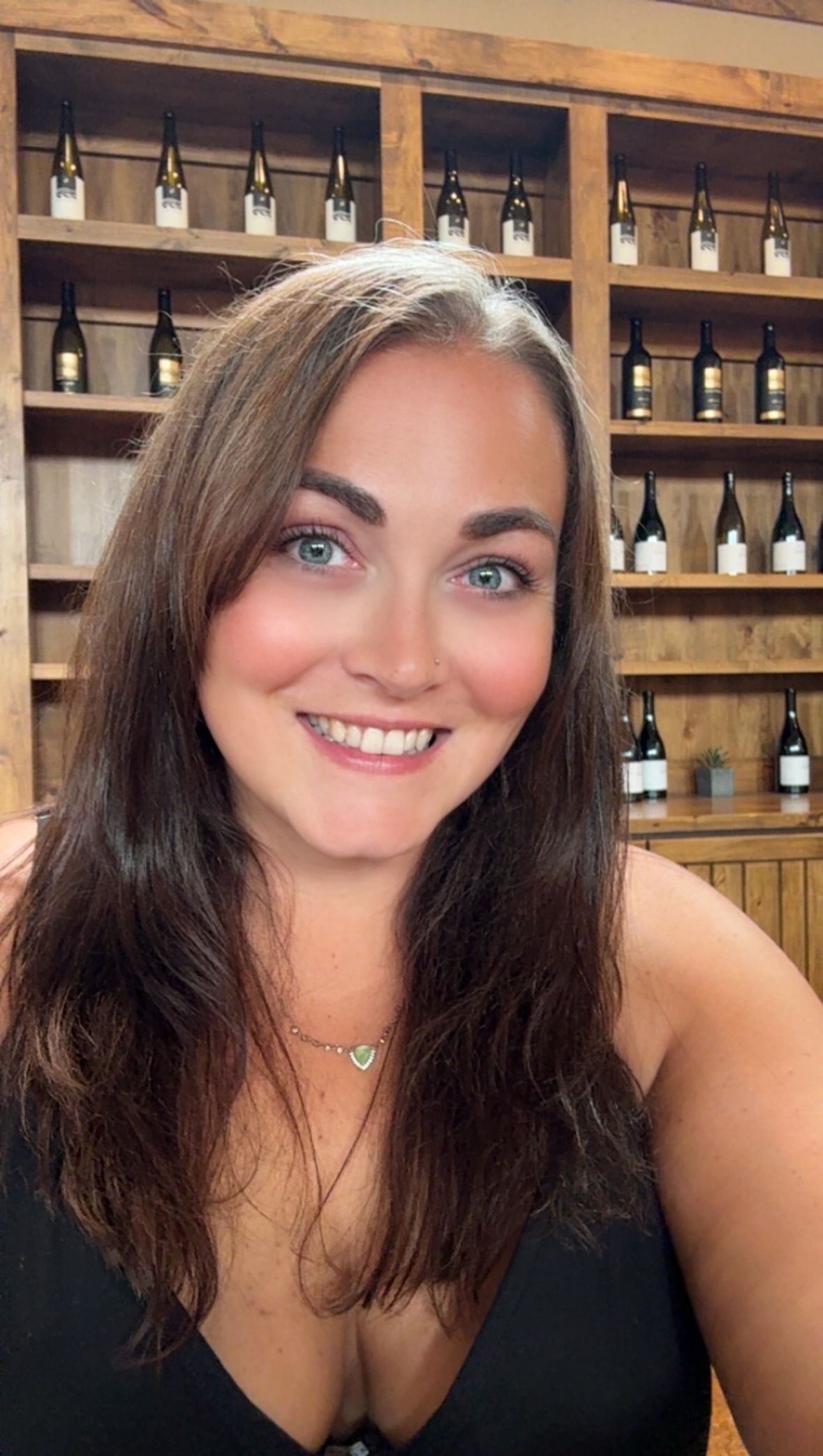 A woman with long brown hair, blue eyes, and a nose piercing is smiling at the camera. She is wearing a black top and a heart-shaped necklace. The background features wooden shelves filled with wine bottles and small potted plants.