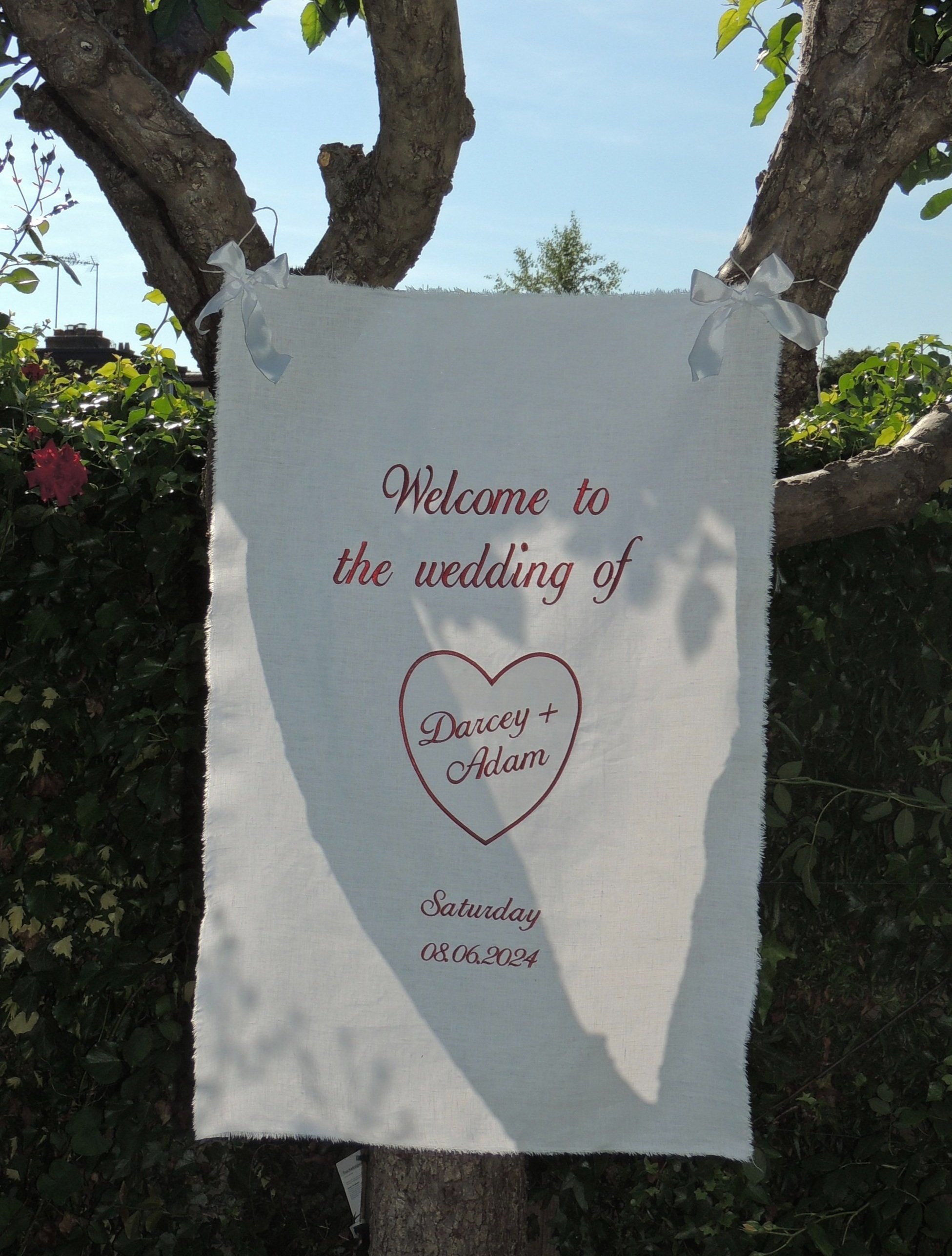 Wedding sign on fabric hanging from a tree reading "Welcome to the wedding of Darcey + Adam, Saturday 08.06.2024."