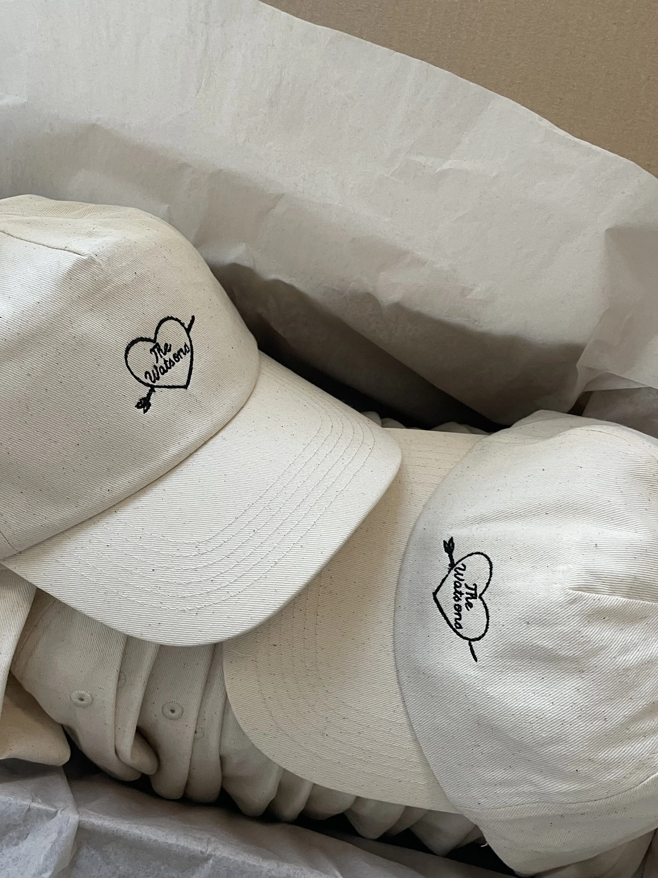 A white baseball cap and a white baseball with black embroidered text that reads 'The Wallets' inside a sleeve or box with white tissue paper.
