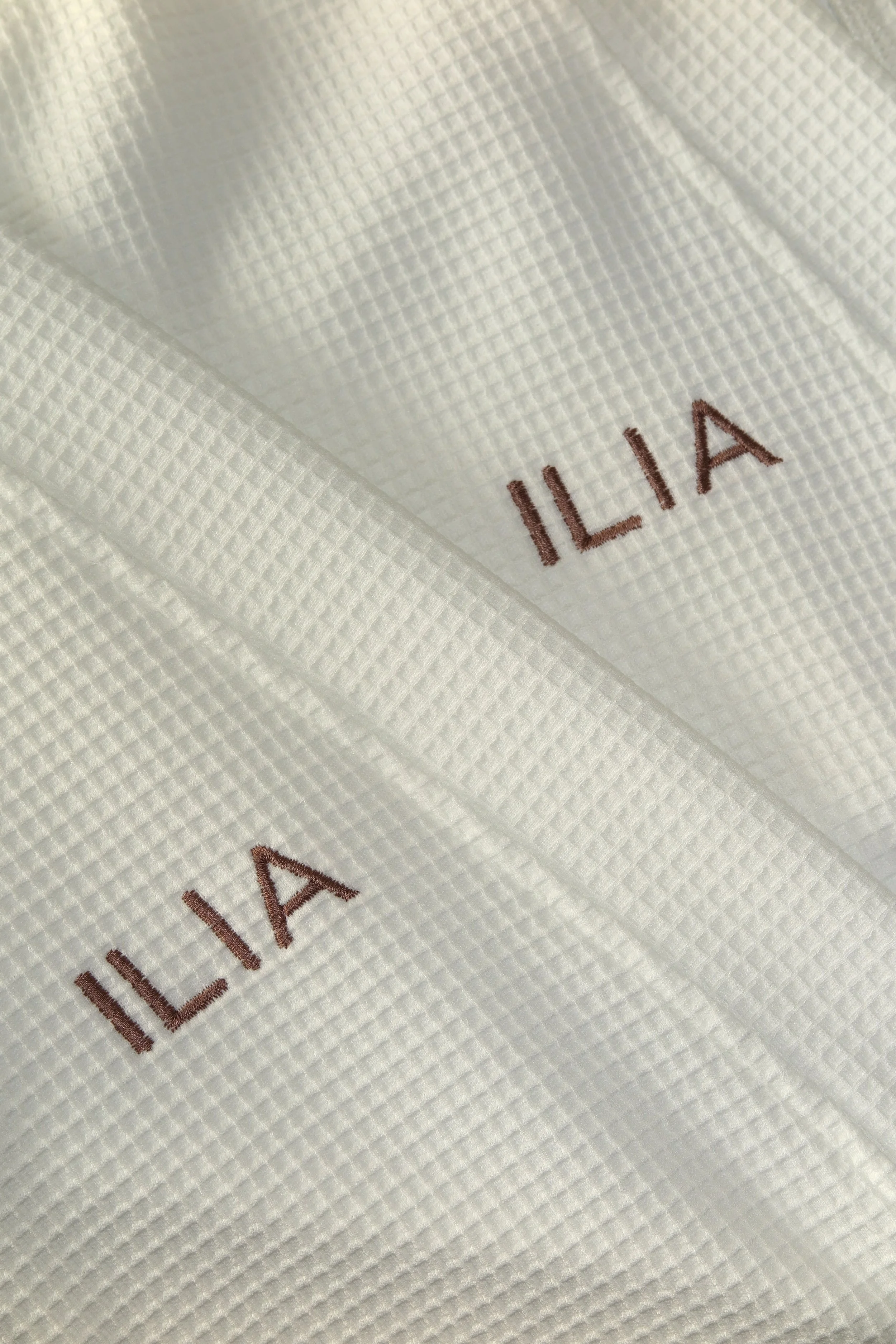 Close-up of white waffle-textured fabric with the word 'ILIA' embroidered in brown lettering.
