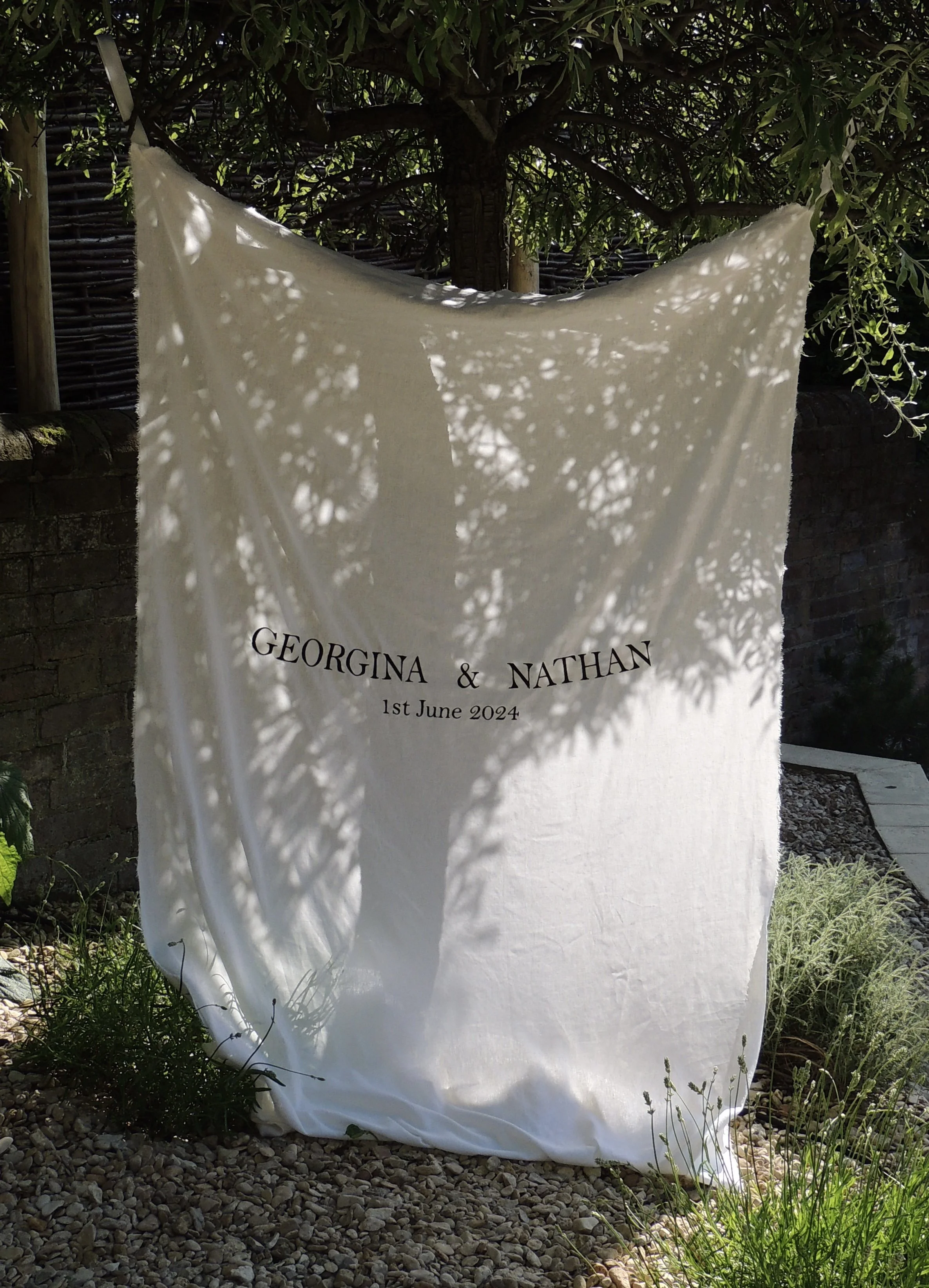 A white cloth with "Georgina & Nathan, 1st June 2024" printed on it, hanging outdoors among leafy trees with shadows cast on its surface.