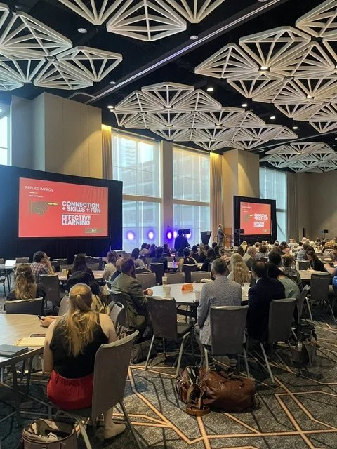 A photo of Malarky Comedy's conference session, with people sitting at round tables facing a podium with two red screens on either side