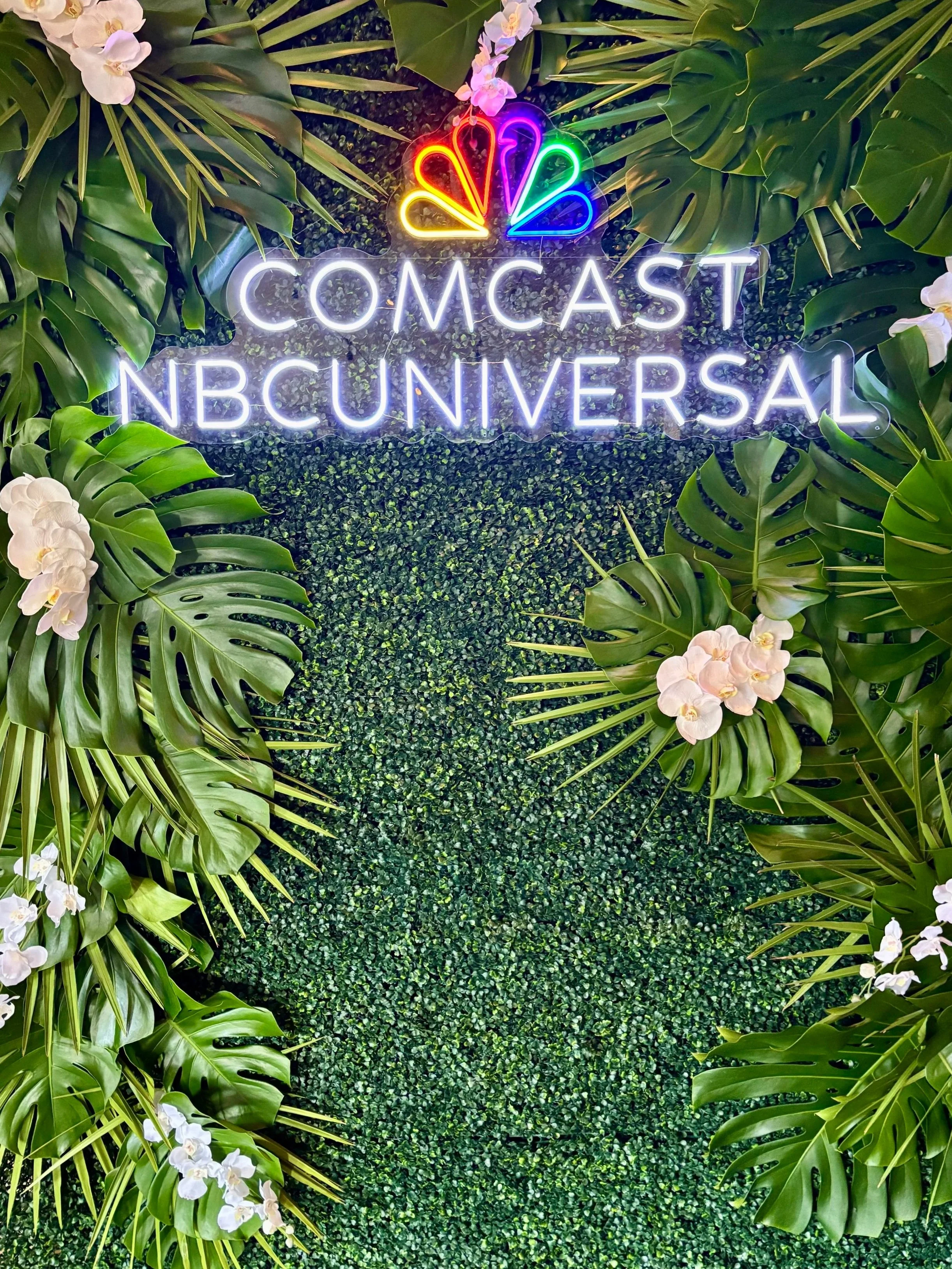 Neon sign displaying the logos of Comcast and NBCUniversal, set against a green leafy backdrop with white orchids.