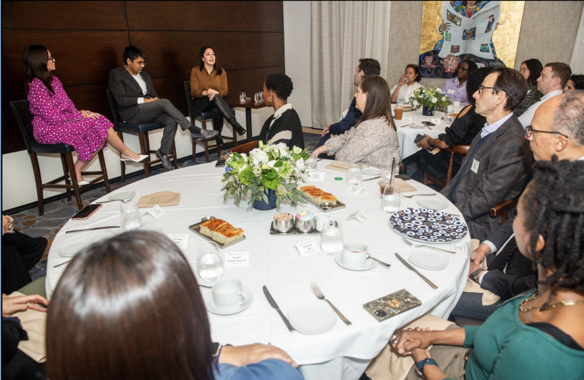 Group of professionals seated around a round table during a discussion at a conference or event with food, drinks, and flowers on the table.