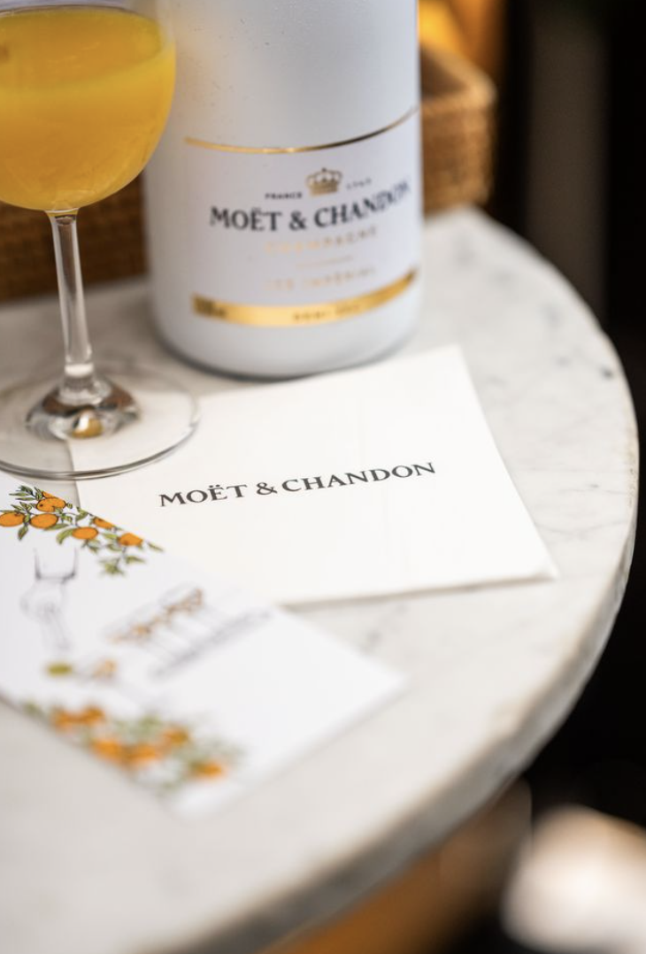 A glass of yellow-orange mimosa on a white marble table with Moët & Chandon champagne packaging and a branded napkin.