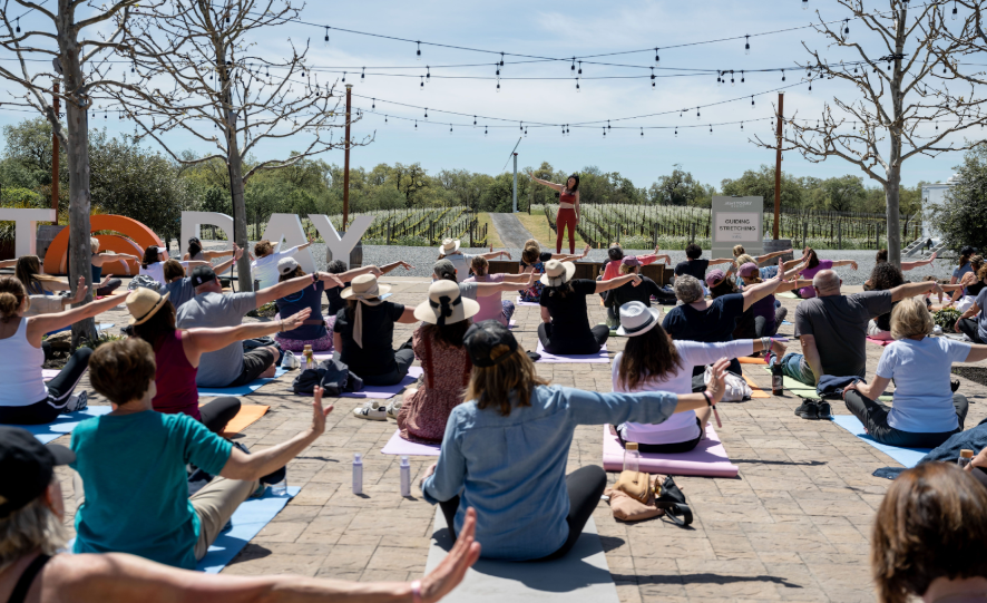 Group of people participating in outdoor yoga class on a sunny day, seated on yoga mats with arms outstretched, facing a instructor on a stage, with string lights overhead and trees in the background.