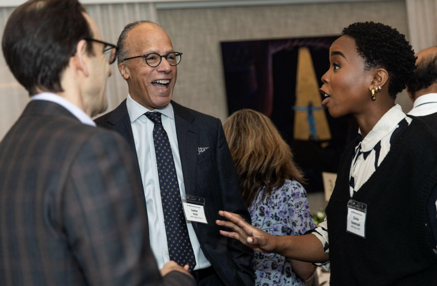 Three people engaging in a conversation at a professional event. The man in the center is smiling warmly, wearing glasses and a suit, while the woman on the right is speaking and gesturing with her hand. The man on the left is partially visible, dressed in a plaid suit.