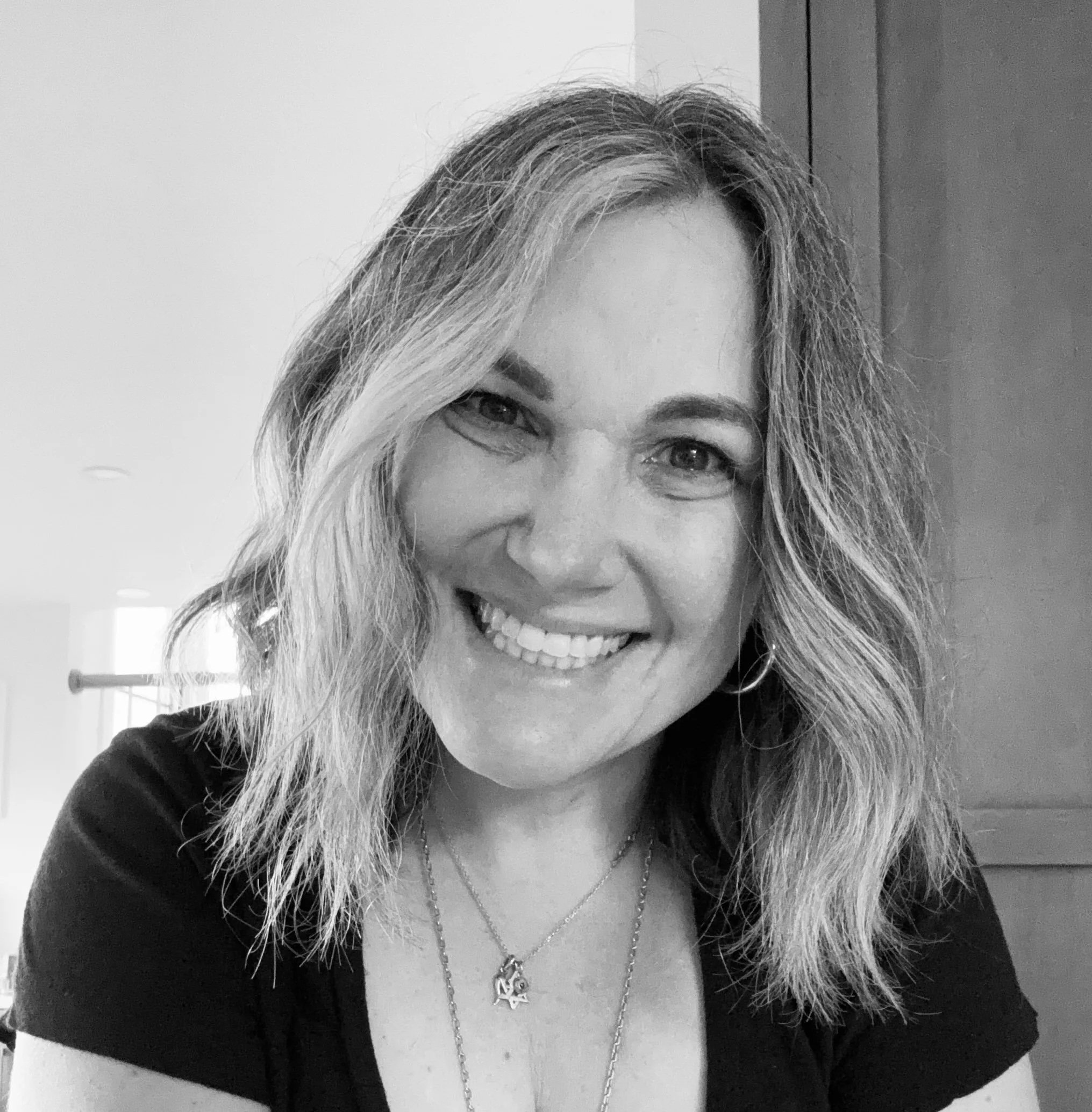 A smiling woman with wavy blonde hair, wearing a necklace and hoop earrings, in a black and white photo.
