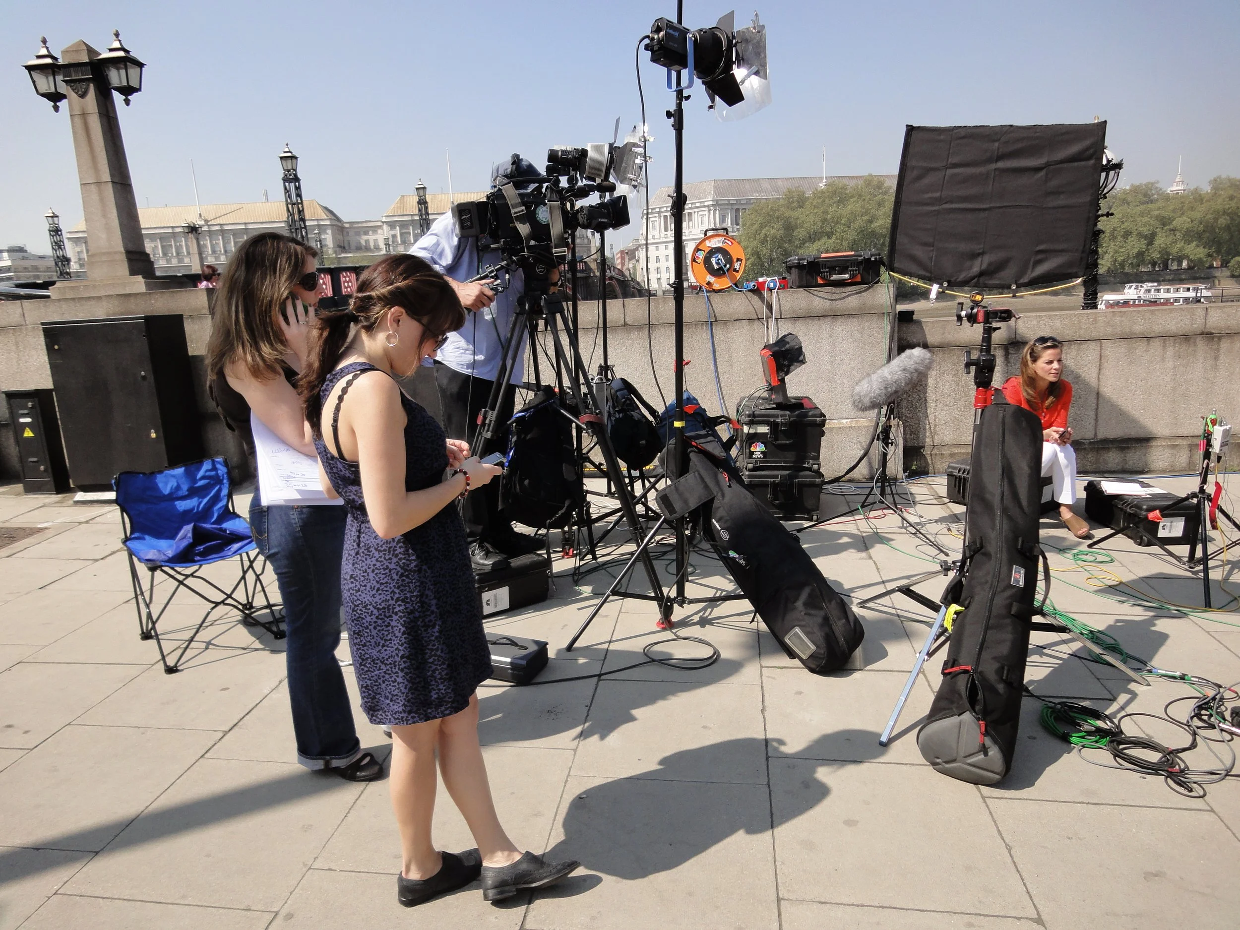 Filming crew setting up cameras and equipment for an outdoor interview with a woman sitting on a small stool in a city park or promenade.