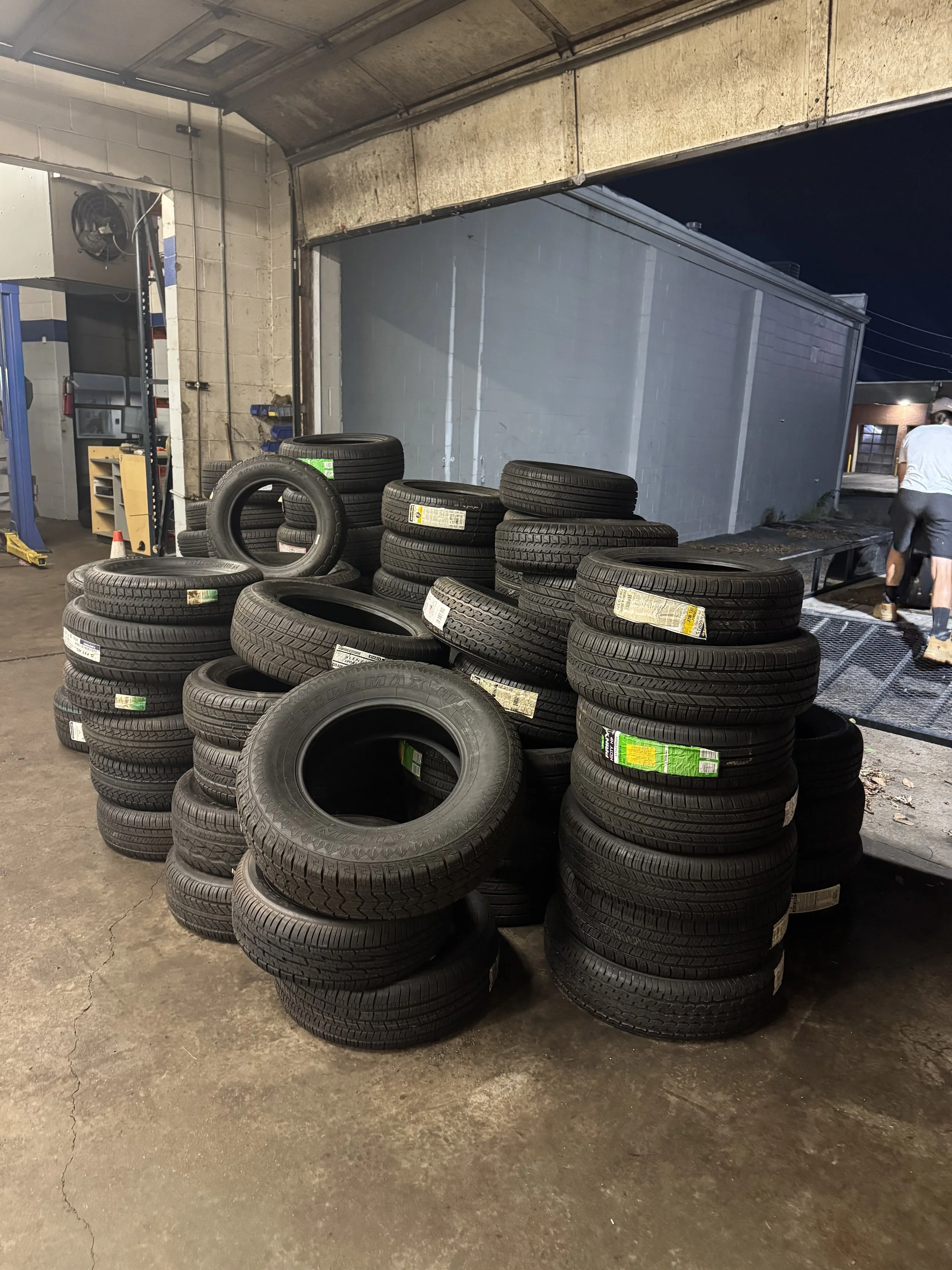 Stacked tires inside a garage or workshop, with some tires on the ground and some on top of each other, near a garage door, with tools and equipment visible in the background.