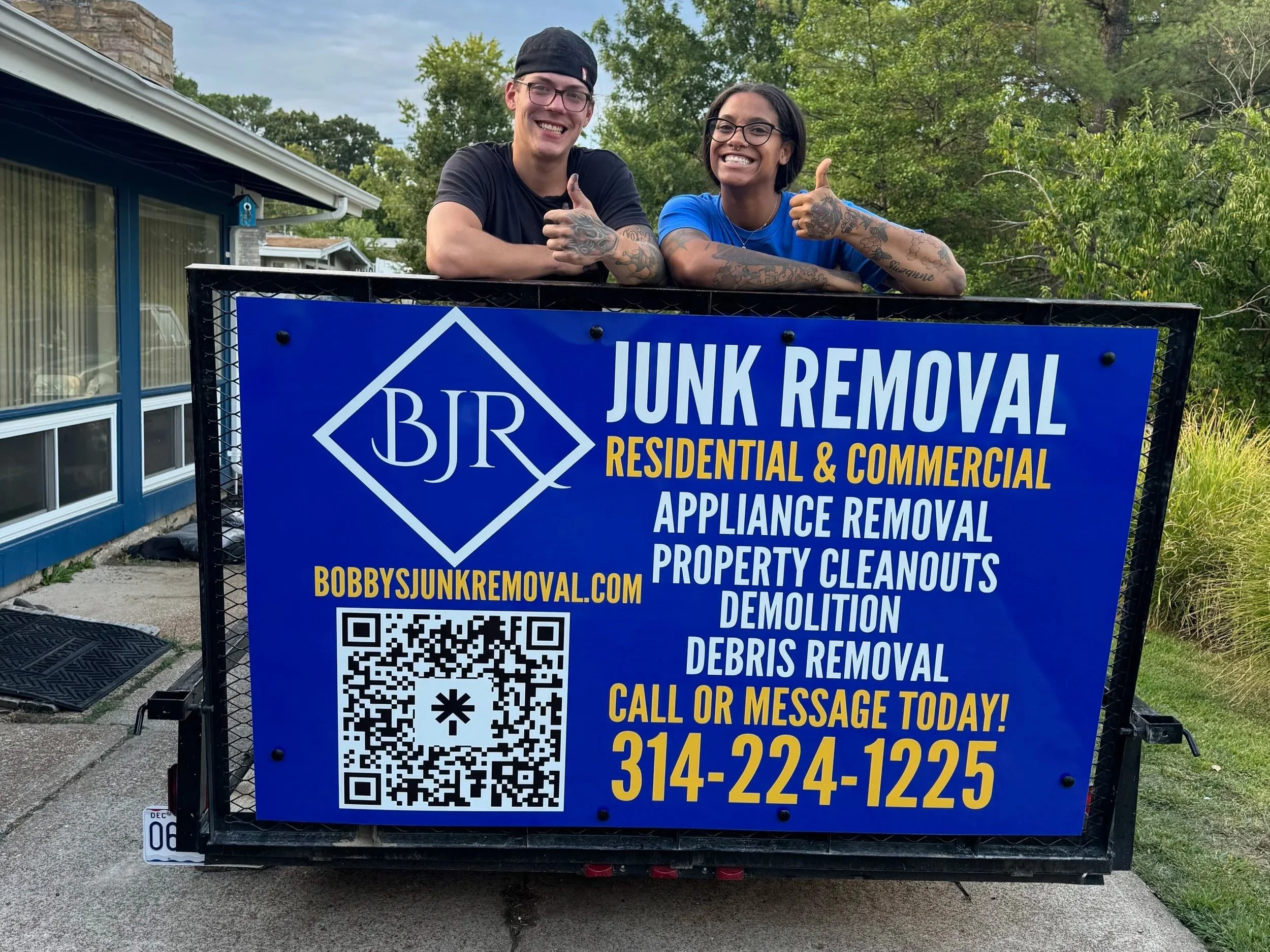 Two smiling people, one man and one woman, leaning on a trailer sign for a junk removal business. The man is wearing a black shirt and cap, and the woman is wearing a blue shirt with glasses. The sign displays the business name 'BJR Junk Removal' and services like appliance removal, property cleanouts, demolition, and debris removal. The sign includes a phone number and website, with a QR code.