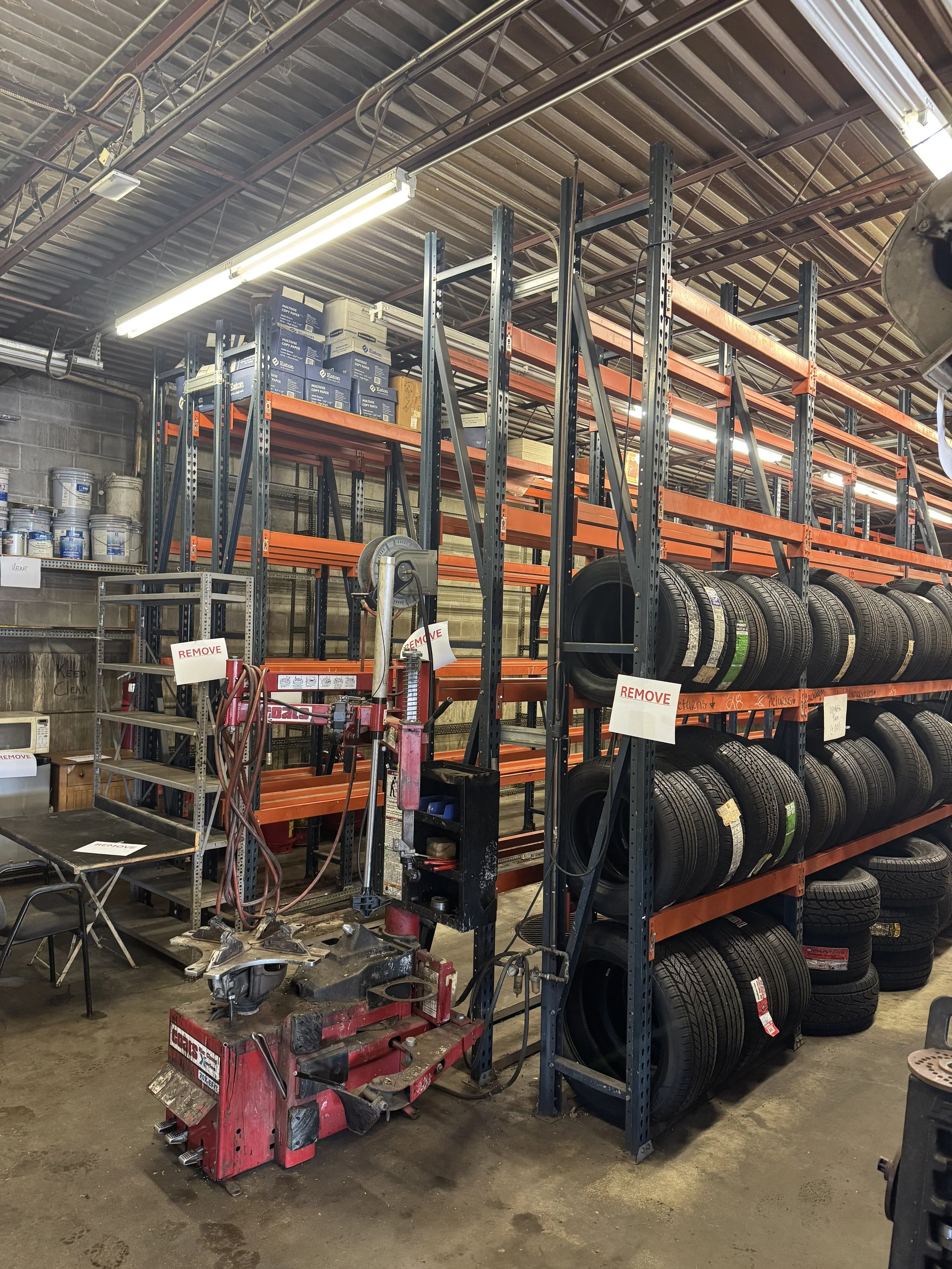 Storage warehouse with car tires on orange metal shelves, a tire-changing machine, and assorted supplies, including paint buckets and a microwave.